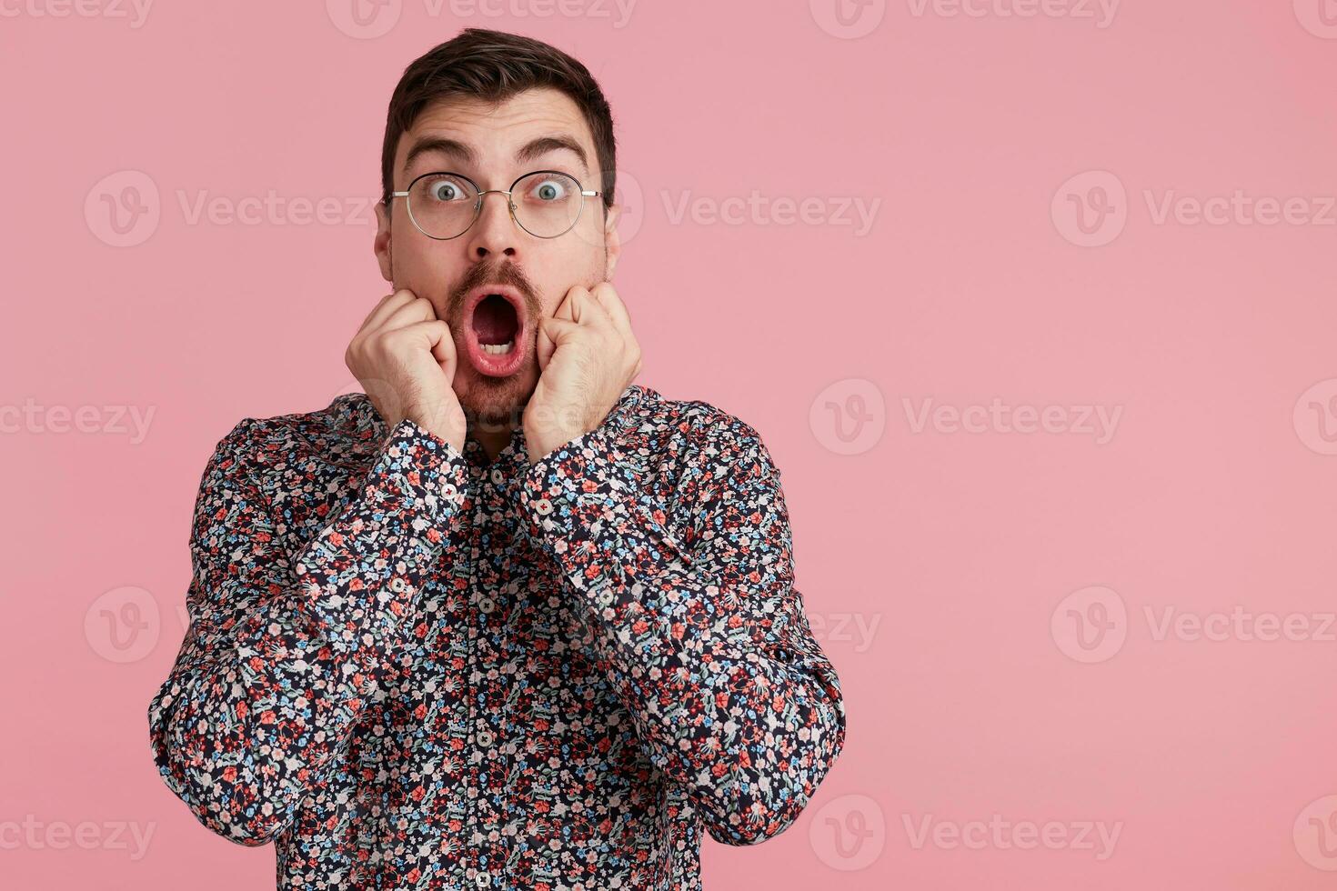 Studio shot of young screaming bearded man in glasses, wearing in colorful shirt, heard the shocking news, with wide open mouth. Isolated over pink background with copy space on the right side. photo