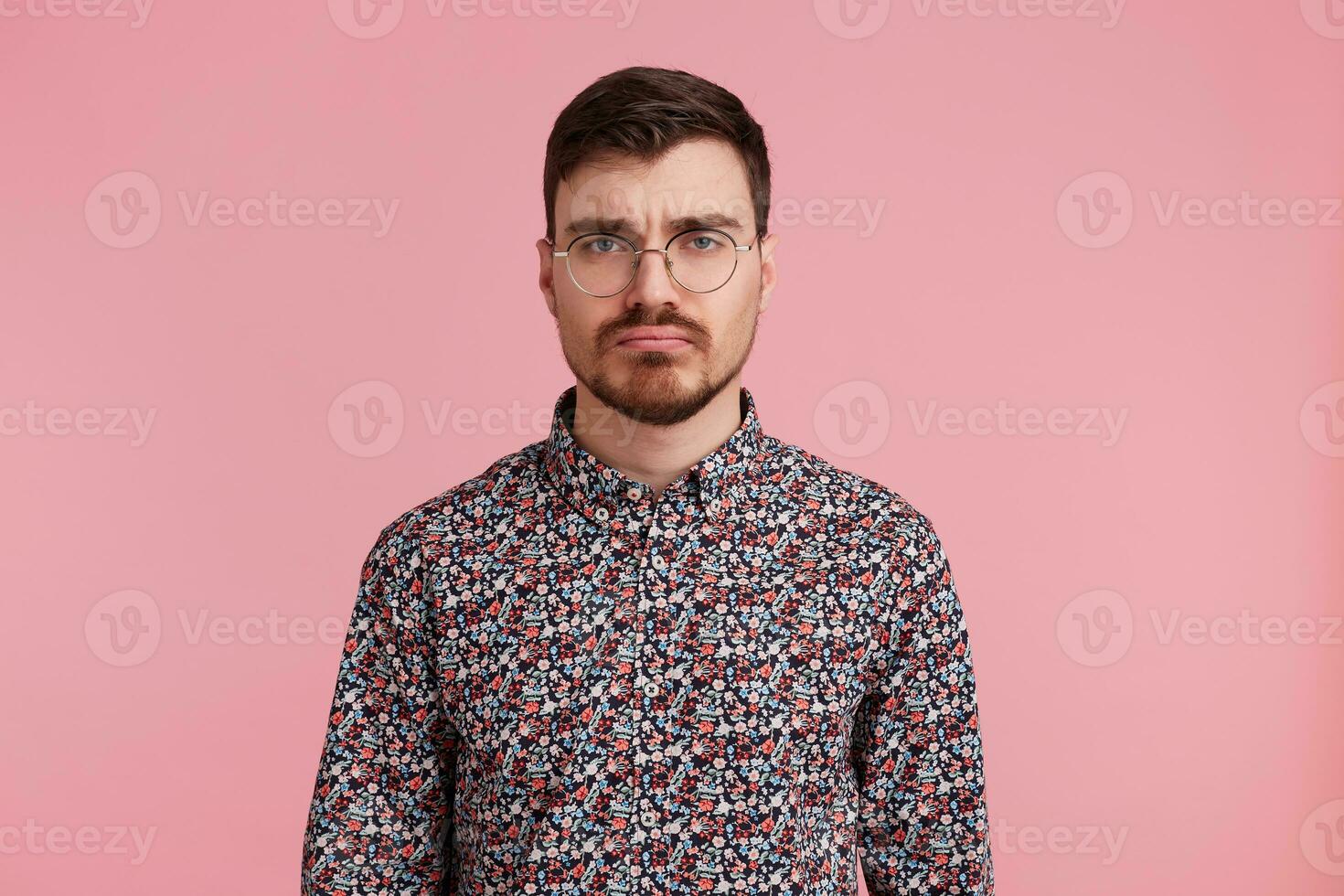 estudio Disparo de triste joven barbado hombre en anteojos, vistiendo en un vistoso camisa, aislado terminado rosado antecedentes. personas y emociones concepto. foto