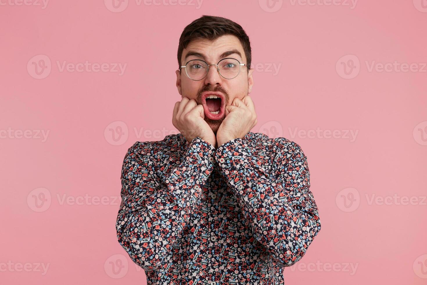 Studio shot of young bearded man in glasses, with wide open mouth, bites finger nails, scared of going to dentist. Wearing in colorful shirt, isolated over pink background. photo