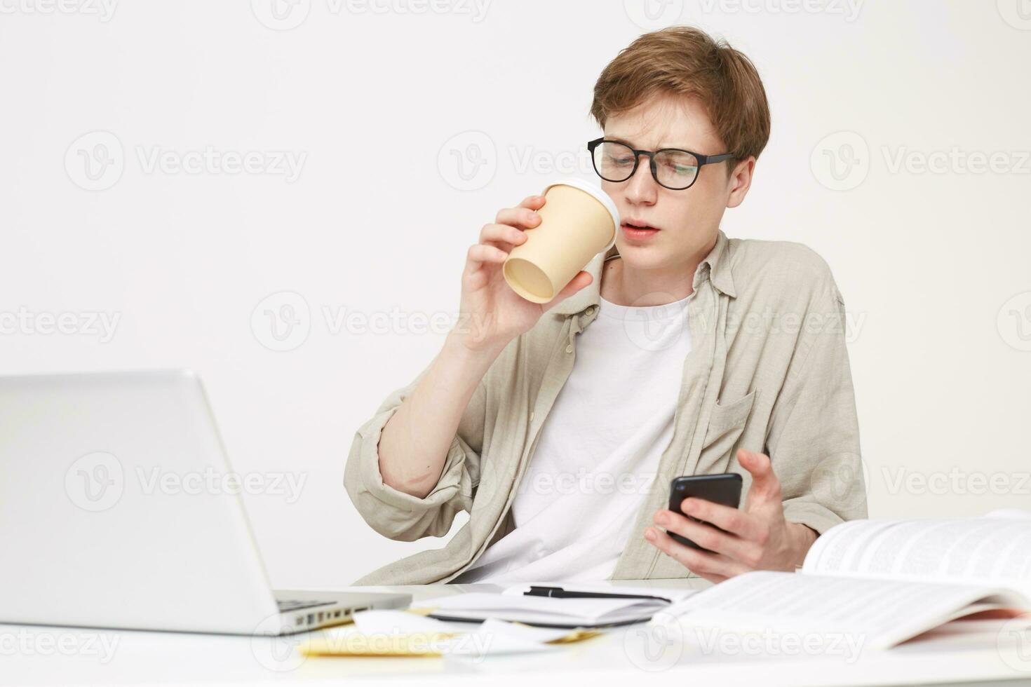 Young man is sitting at the table, drinking coffee, holding a phone in his hand, a notebook pen is on the table and laptop opened,looks on smart phone photo