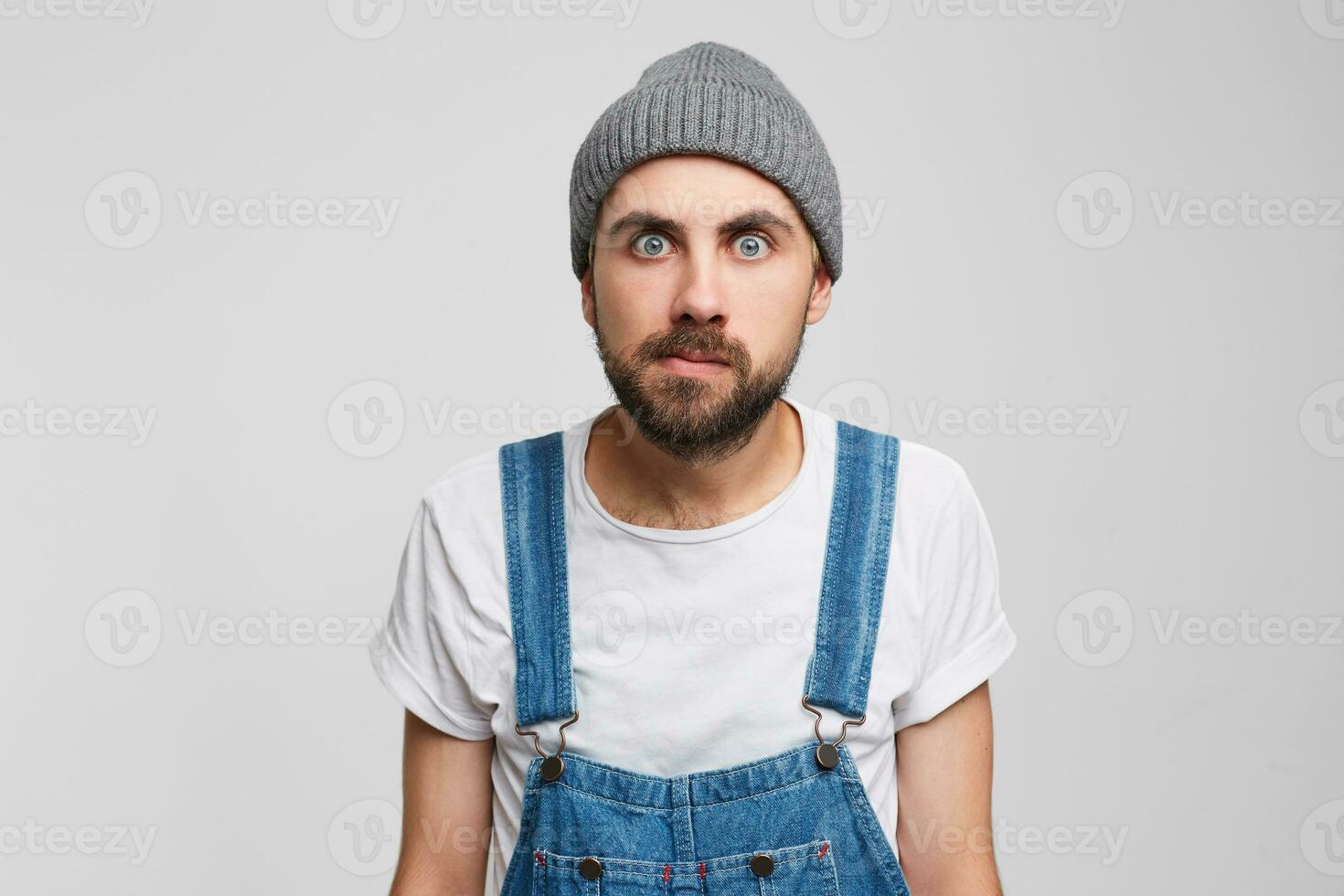 Young man did not slept for a long time, tired, zombie eyes, wide-open eyes, staring, one corner of the lips is pursed, dressed in grey hat and overall, isolated over white background photo