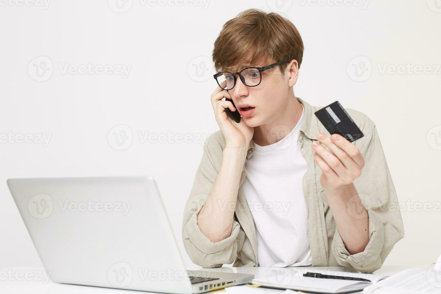 Studio shot of young man sits at a table, holds a credit card in his hands, talking to a bank manager confused, frustrated face expression, a notebook pen and an open laptop are on the table photo
