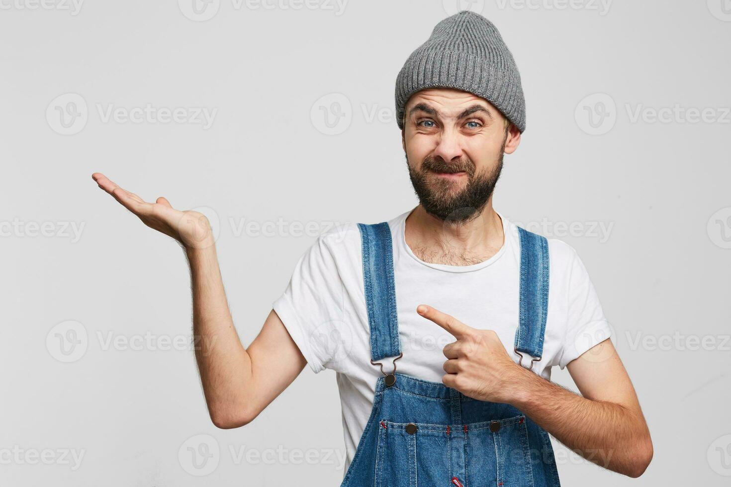 A man with a displeased squeamish unsatisfied facial expression looks into the camera, with his index finger shows towards to copy space on his palm, a white background photo