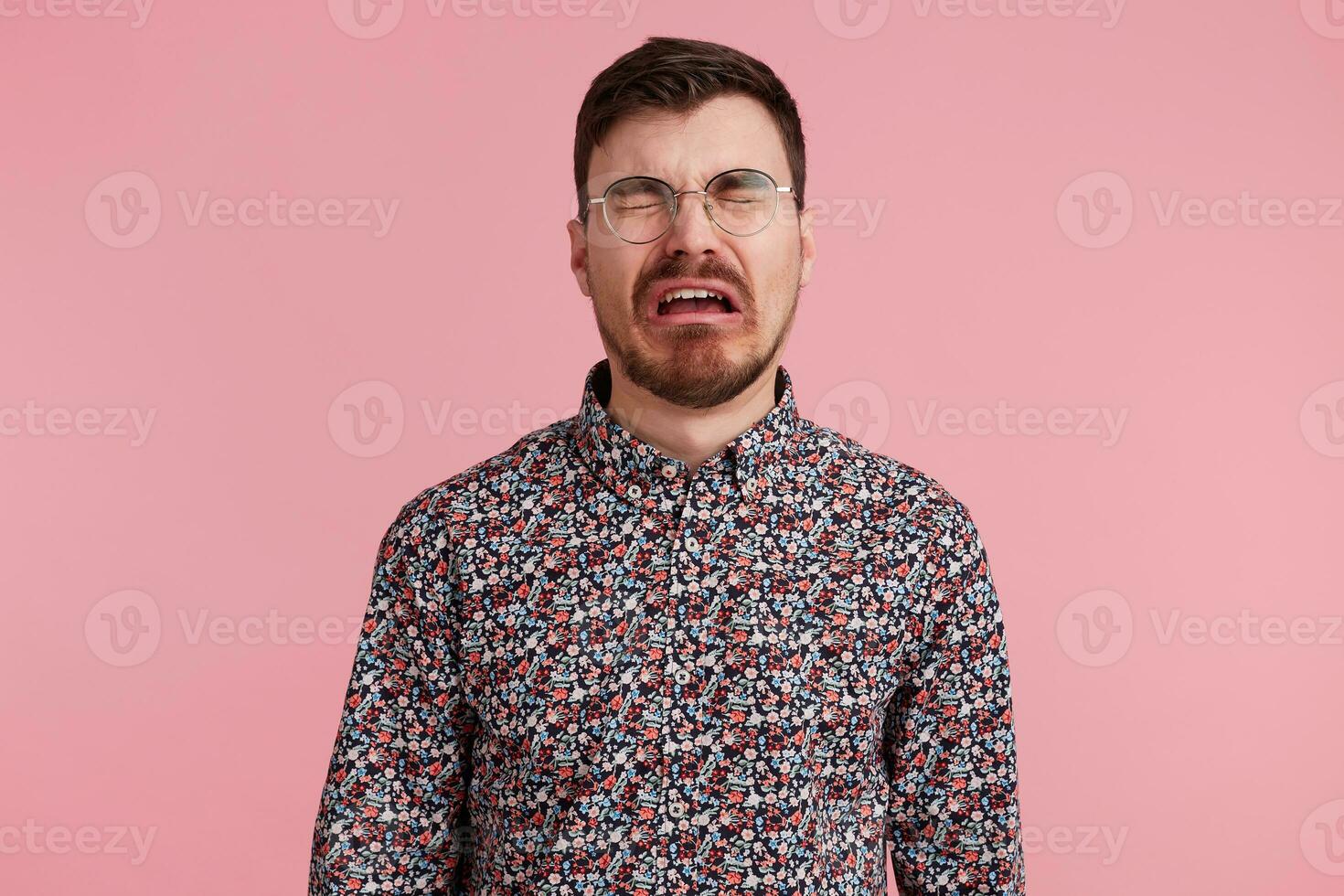 Portrait of a sad crying bearded man in glasses, wearing in colorful shirt, looks unhappy and upset. Isolated over pink background, people and emotions concept. photo