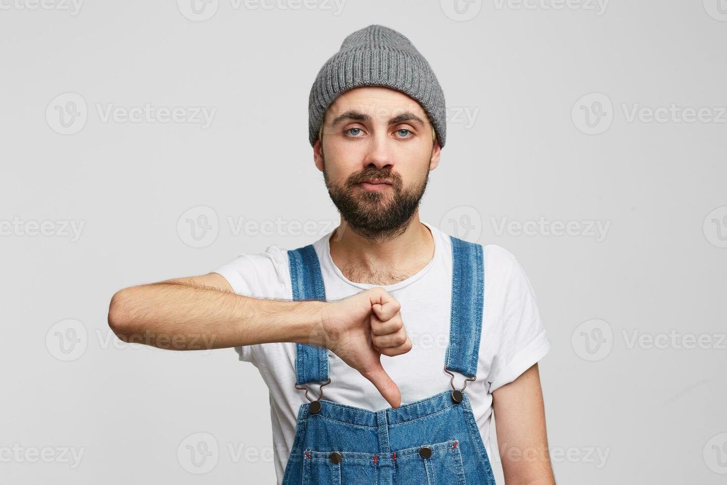 The guy gives a negative assessment, standing against white background with an indifferent unsatisfied facial expression, with his hand shows a thumb down. Problem, bad mood photo