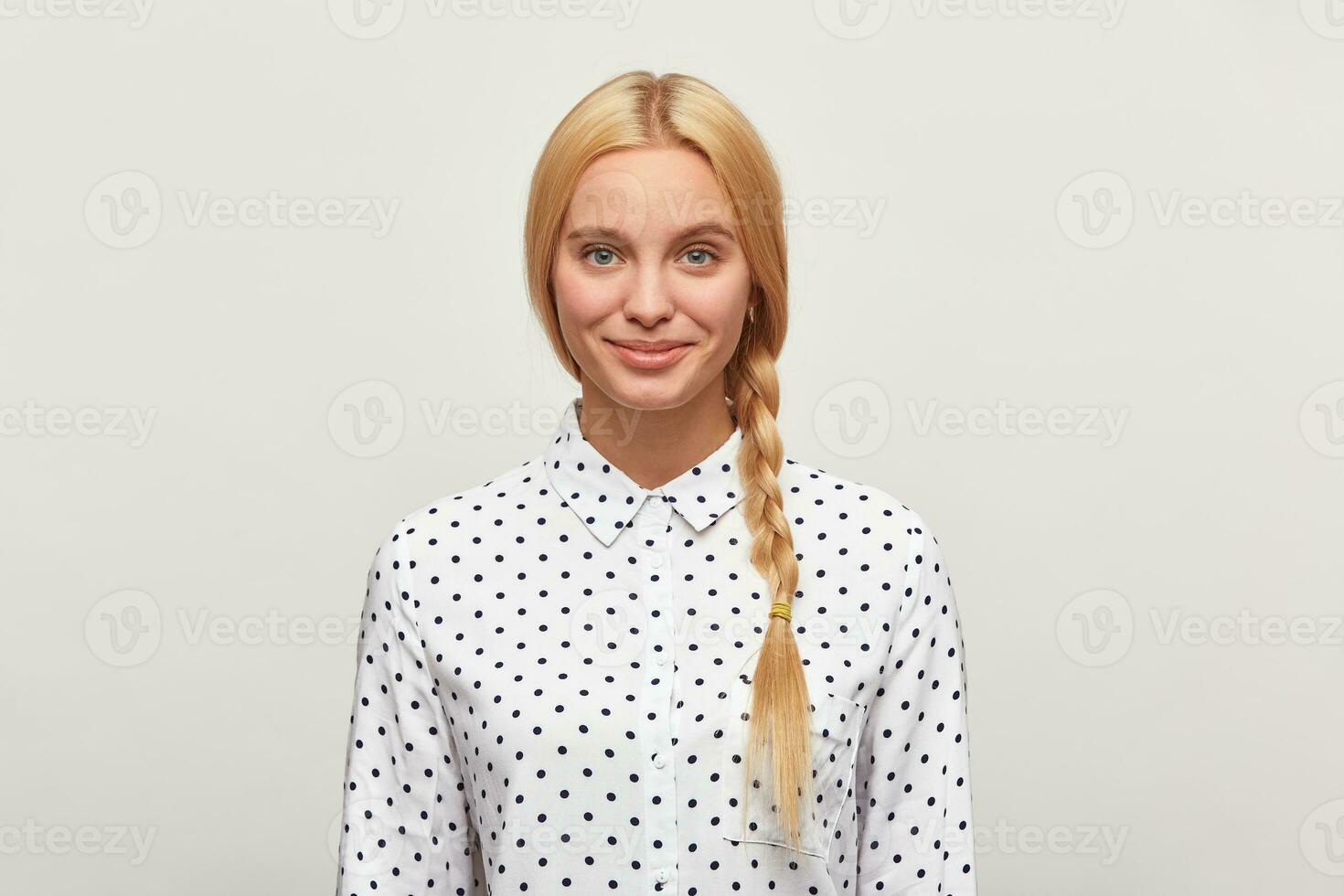 Beautiful diligent blonde woman with blue eyes, braid, feels happy, tenderly smiles, wears white shirt with black polka dots. Modest, excellent student, came to interview. Over white background photo