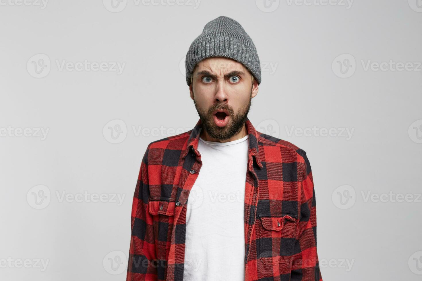 A young man disagrees with the interlocutor's answer and is ready to argue with him, express anger, mouth wide opened, wrathful look. Wears checkered shirt, unshaven, isolated over white background photo