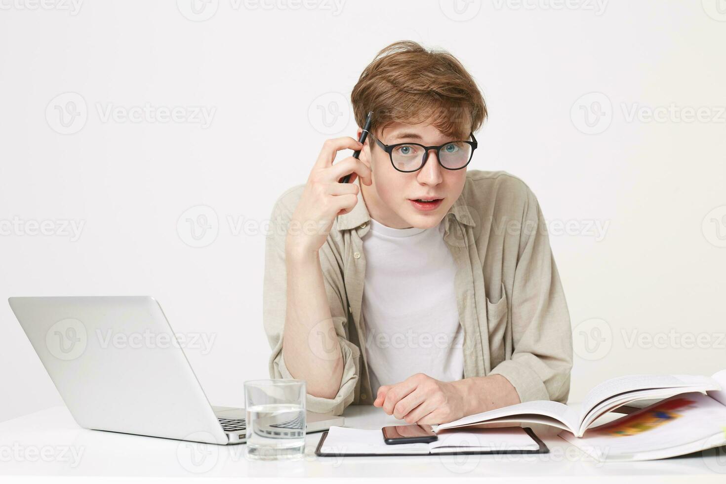 Student is sitting at the table, textbooks and notebooks are laid out in front of him, looking directly at the camera through his glasses, holding a pen near the temple, thinking, pondering the task photo