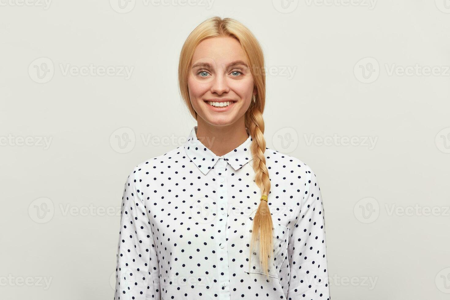 Portrait of a beautiful blonde young girl with natural make-up and well-groomed hair gathered in braid tenderly smiles, wears white shirt with black polka dots, on a white background in the studio. photo