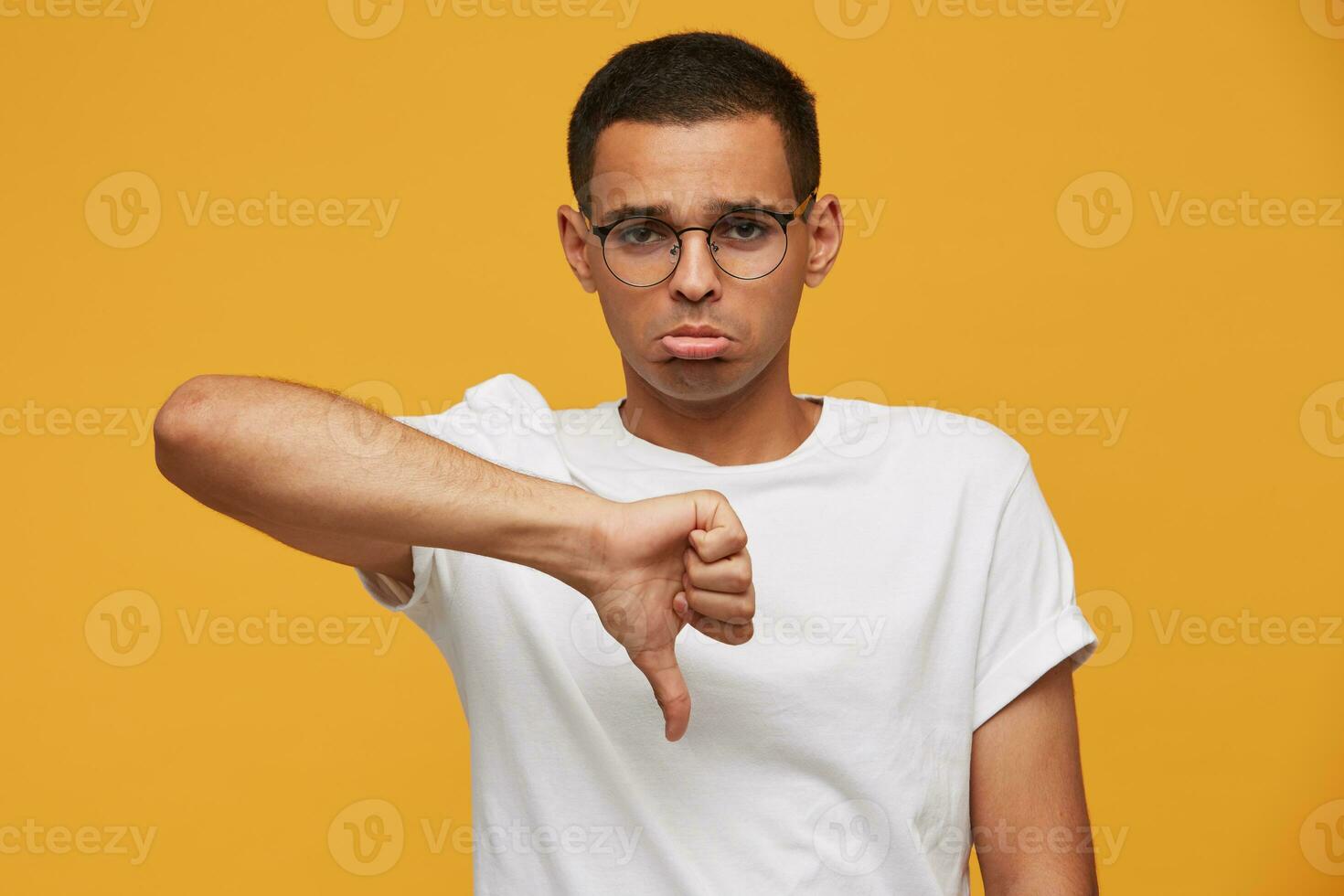 Portrait of young man in glasses looks sad, upset, frustrated, displeased with the result of the game, finger down, lips pout, wears white casual t-shirt, on a yellow background photo