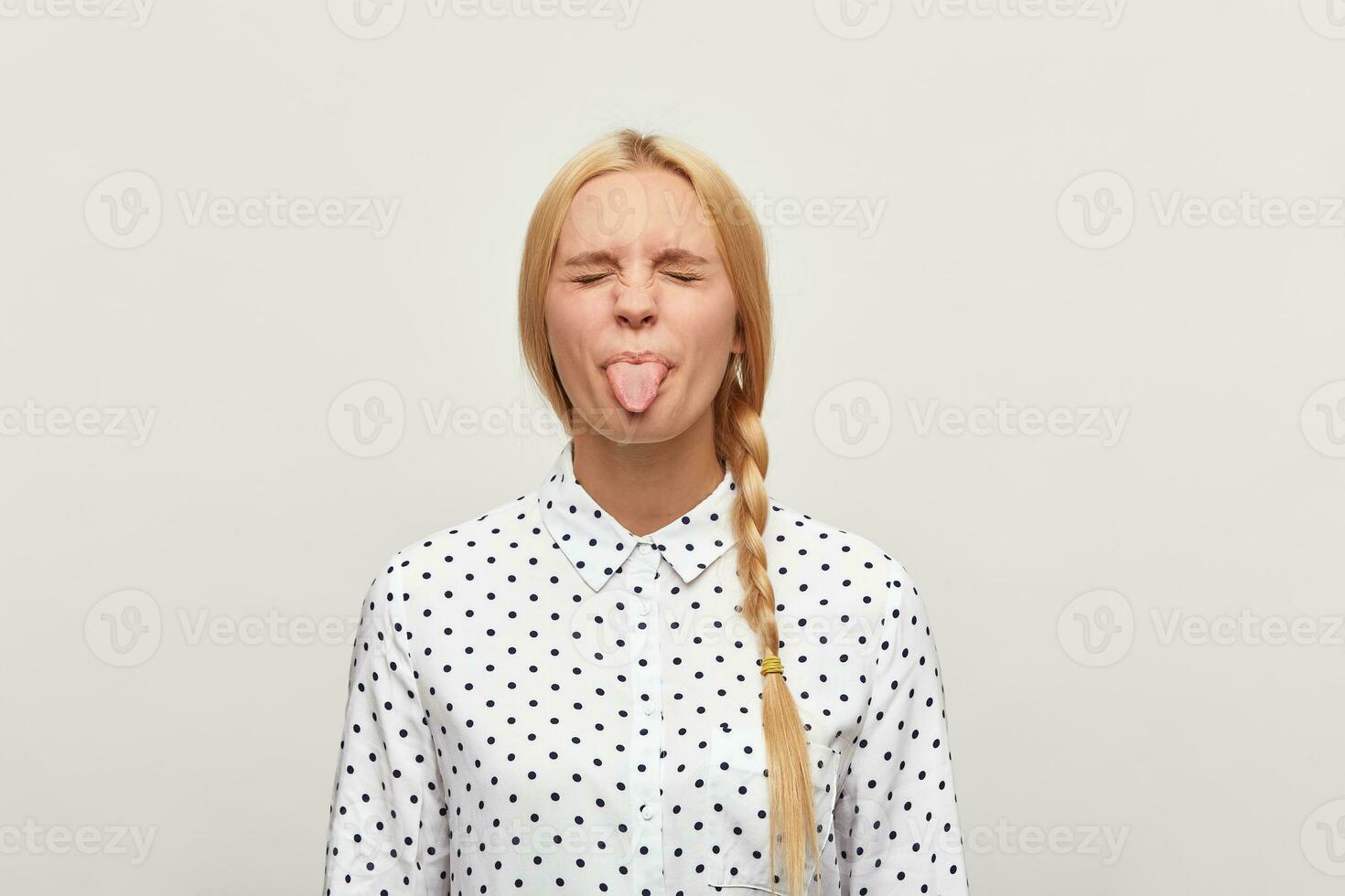 The girl expresses insult childly. Close up of blonde young woman, feels upset, shows tongue eyes closed, wears white shirt with black polka dots isolated over white background photo