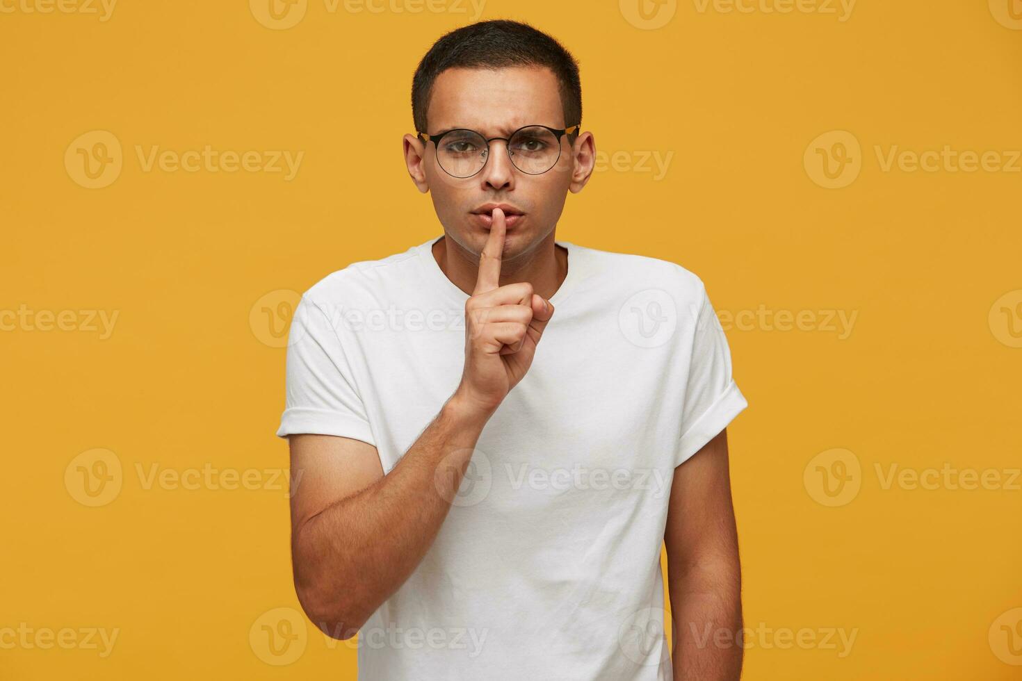 Emotional young man in glasses calls for silence, keep a secret, do not tell anyone, keep quiet, do not make noise, keeps fore finger on lips, demonstrates silence gesture, on a yellow background photo