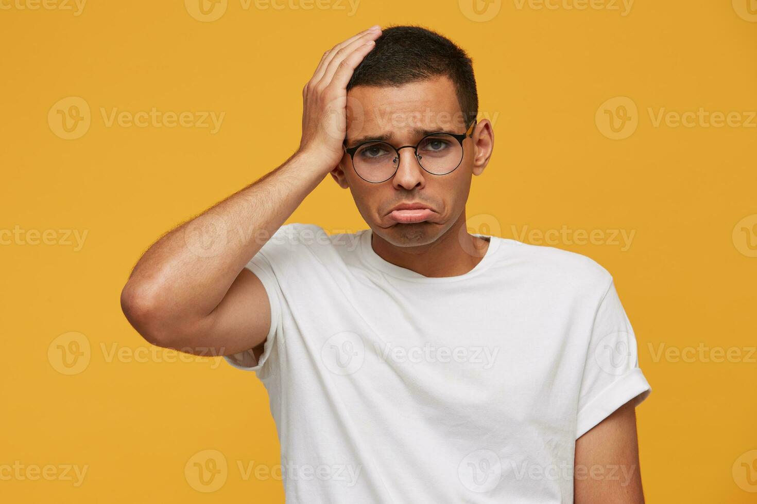 Young man in glasses looks sad, upset, frustrated, displeased with the result of the football match, team play, lips pout, put his hand on head, wears white casual t-shirt, on a yellow background photo