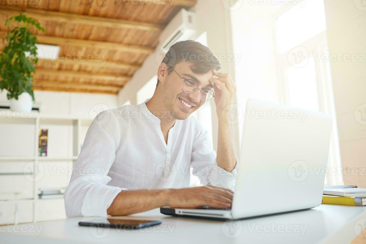 Portrait of happy confident young businessman wears white shirt and glasses using laptop and smartphone sitting at the table, working in office and laughing photo