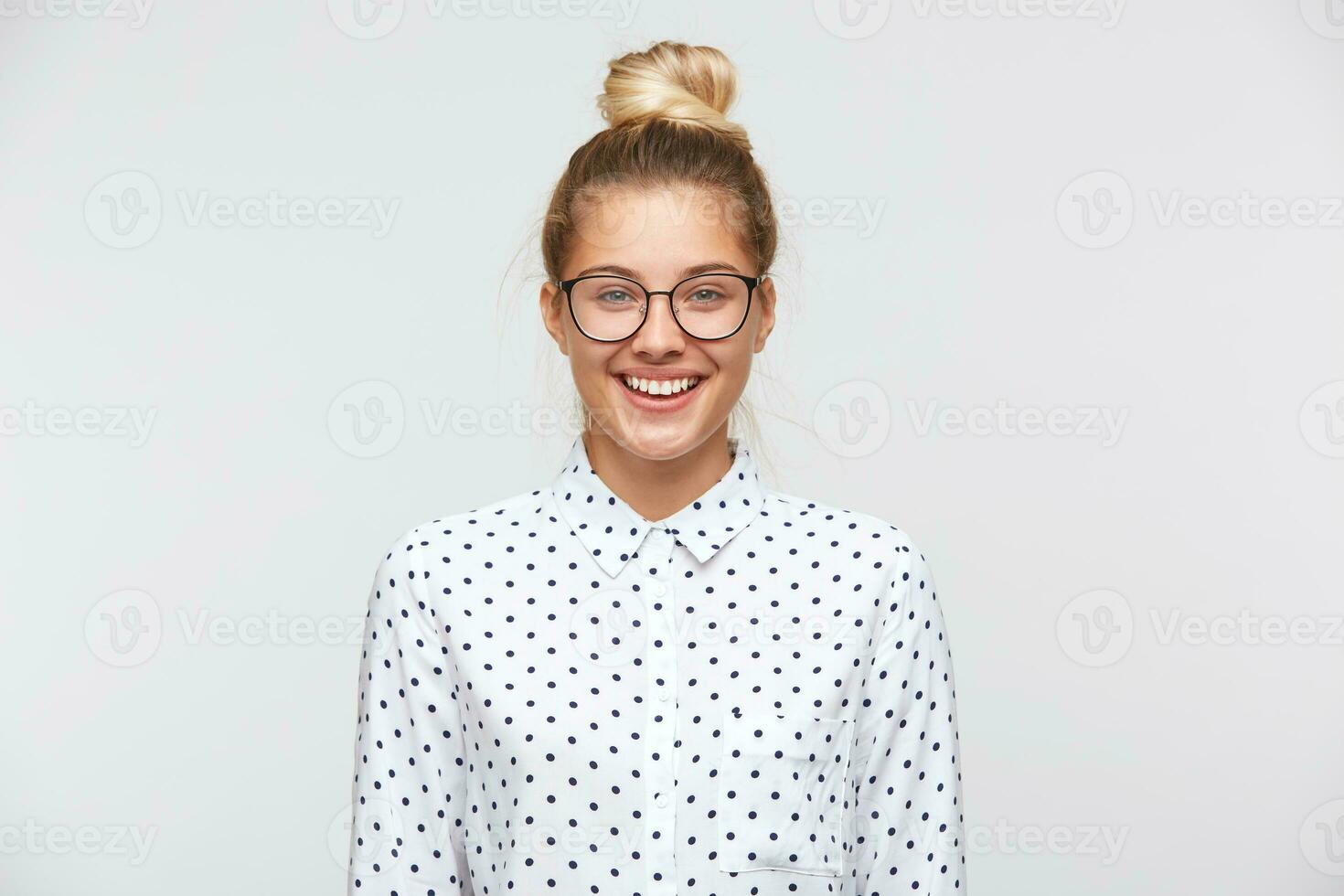 Closeup of smiling pretty young woman with bun wears polka dot shirt and glasses feels happy and confident isolated over white background photo