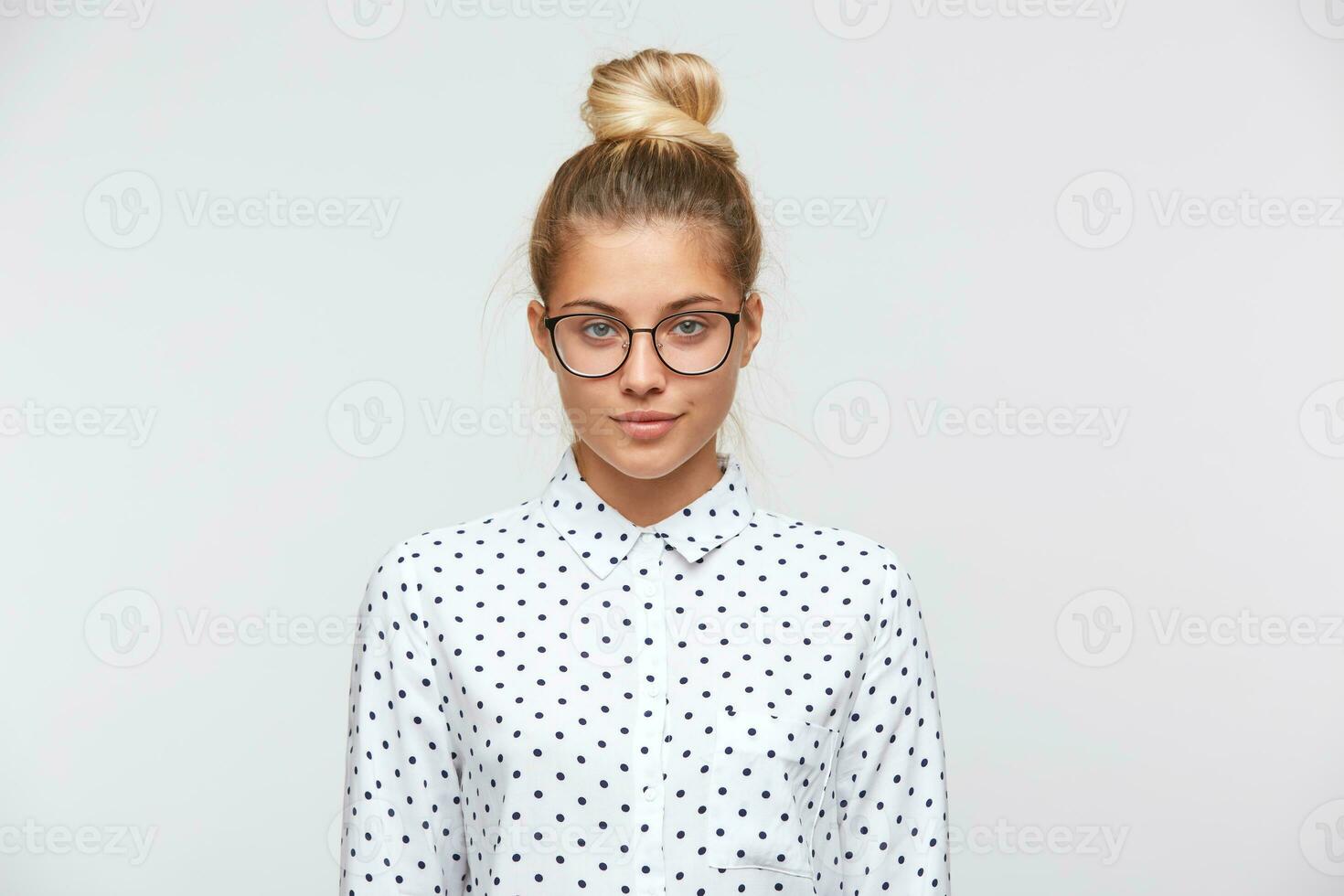 Portrait of confident attractive young woman with bun wears polka dot shirt and spectacles feels serious and looks to the camera isolated over white background photo