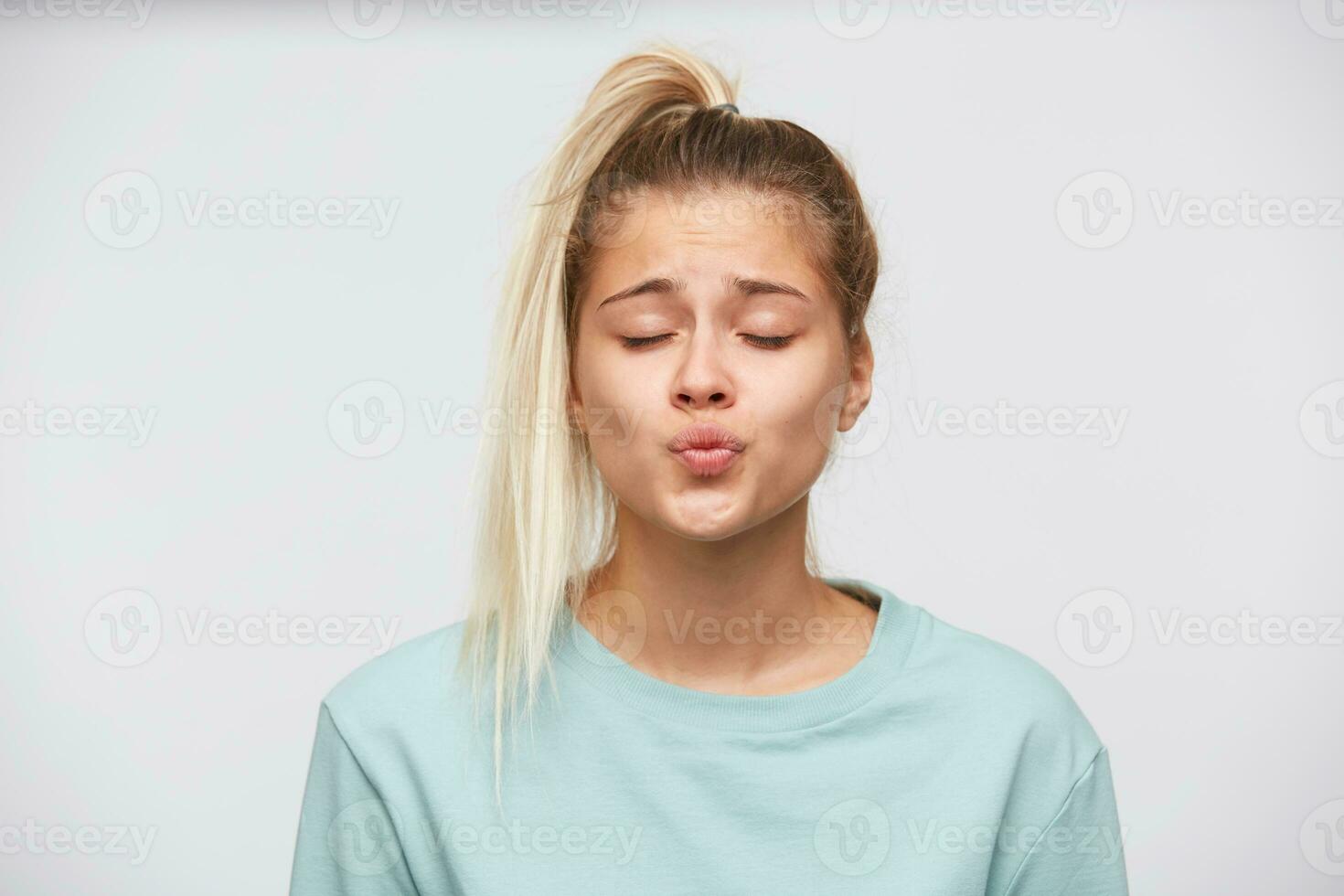 Portrait of happy cute young woman with blonde hair, closed eyes and ponytail wears blue sweatshirt looks playful and kissing isolated over white background photo
