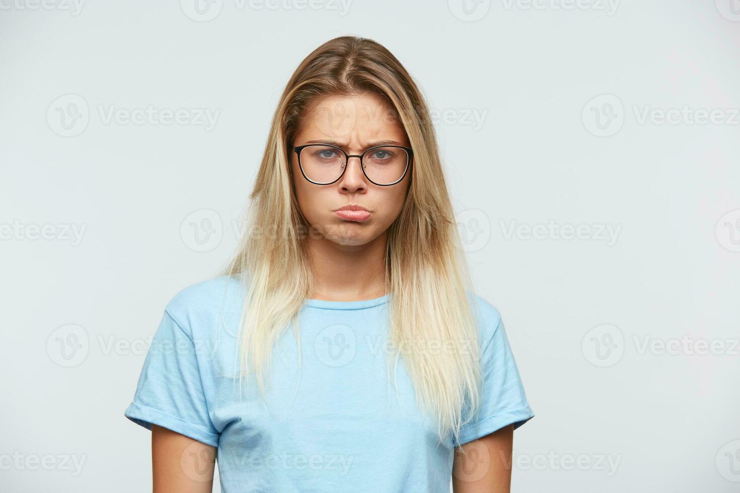 Portrait of unhappy disappointed young woman with blonde hair wears glasses and blue t shirt feels sad and curving lips isolated over white background photo