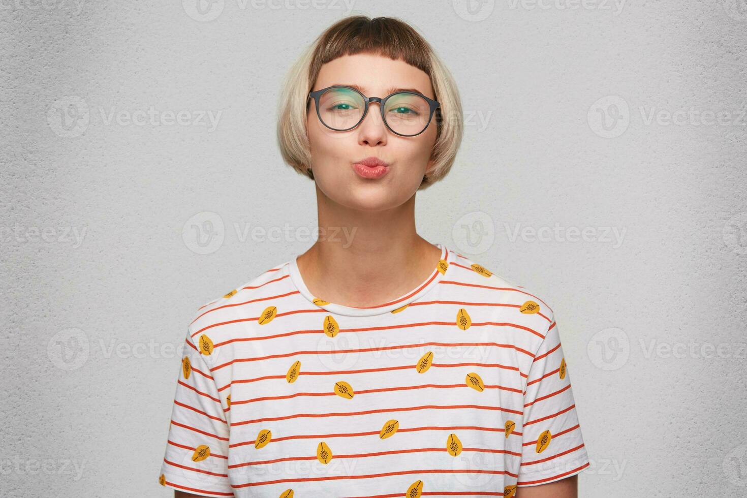 Closeup of happy attractive young woman wears striped t shirt and spectacles looks playful and pretending kissing by lips isolated over white background photo