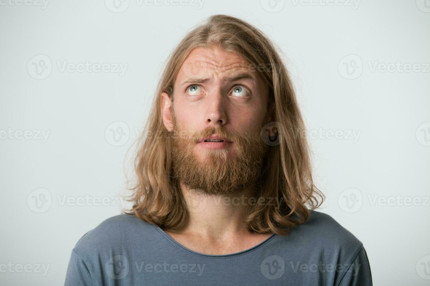 Closeup of pensive handsome young man with beard and blonde long hair wears gray t shirt feels puzzled and thinking isolated over white background photo