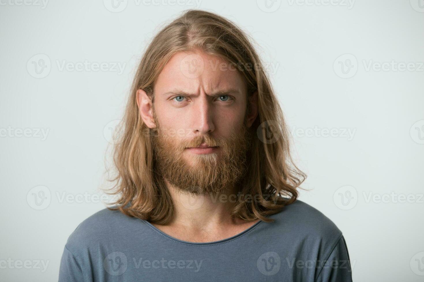 Portrait of angry thoughtful bearded young man with blonde long hair wears gray t shirt looks irritated and serious isolated over white background photo