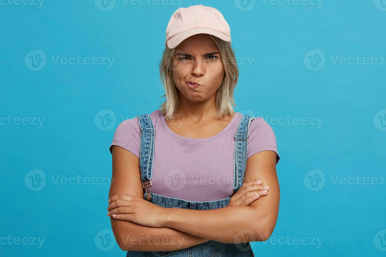 Closeup of unhappy upset blonde young woman wears pink cap, violet t shirt and denim jumpsuit looks displeased and keeps arms crossed isolated over blue background photo