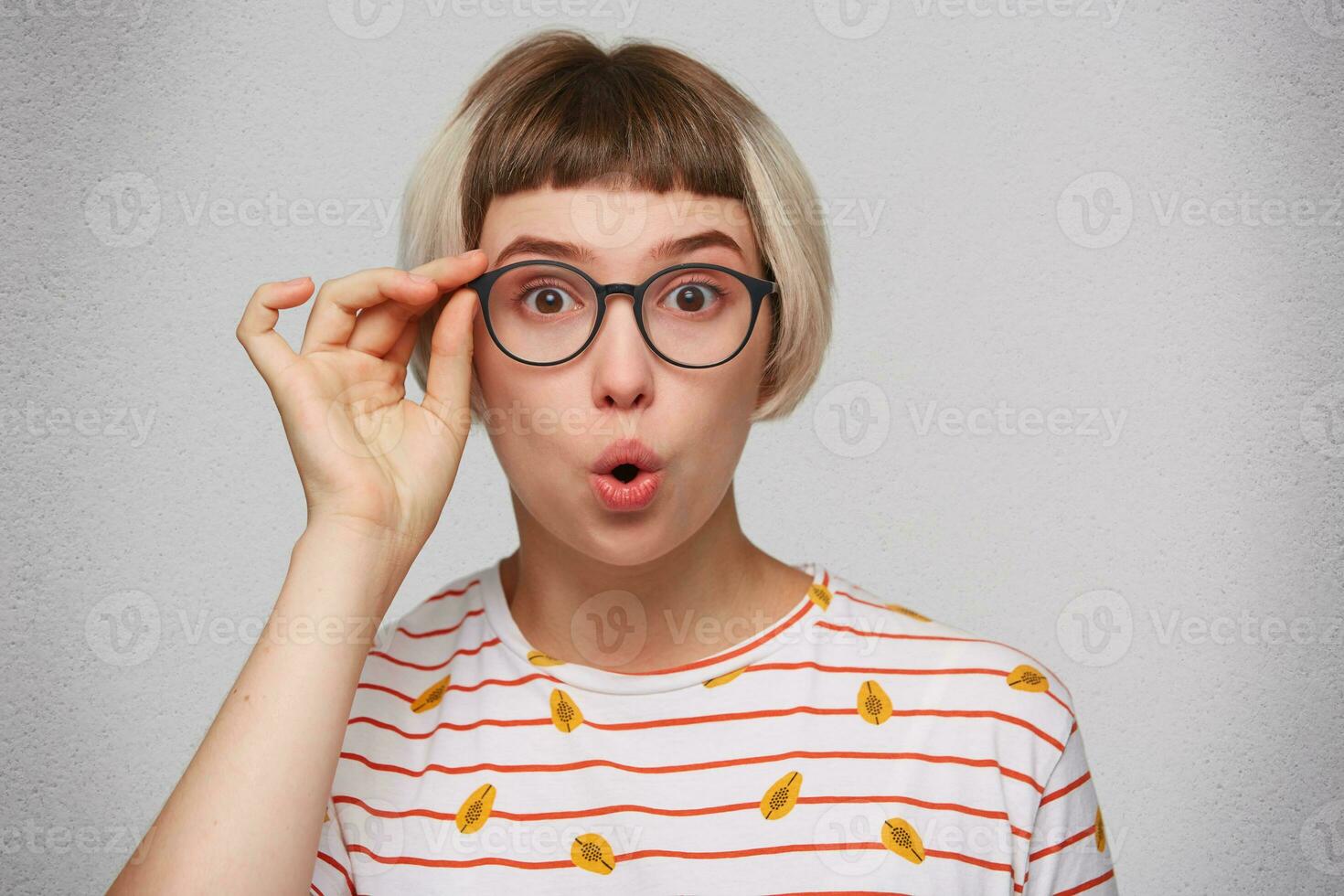 Portrait of shocked stunned young woman wears striped t shirt and glasses feels amazed and looks directly in camera isolated over white background photo