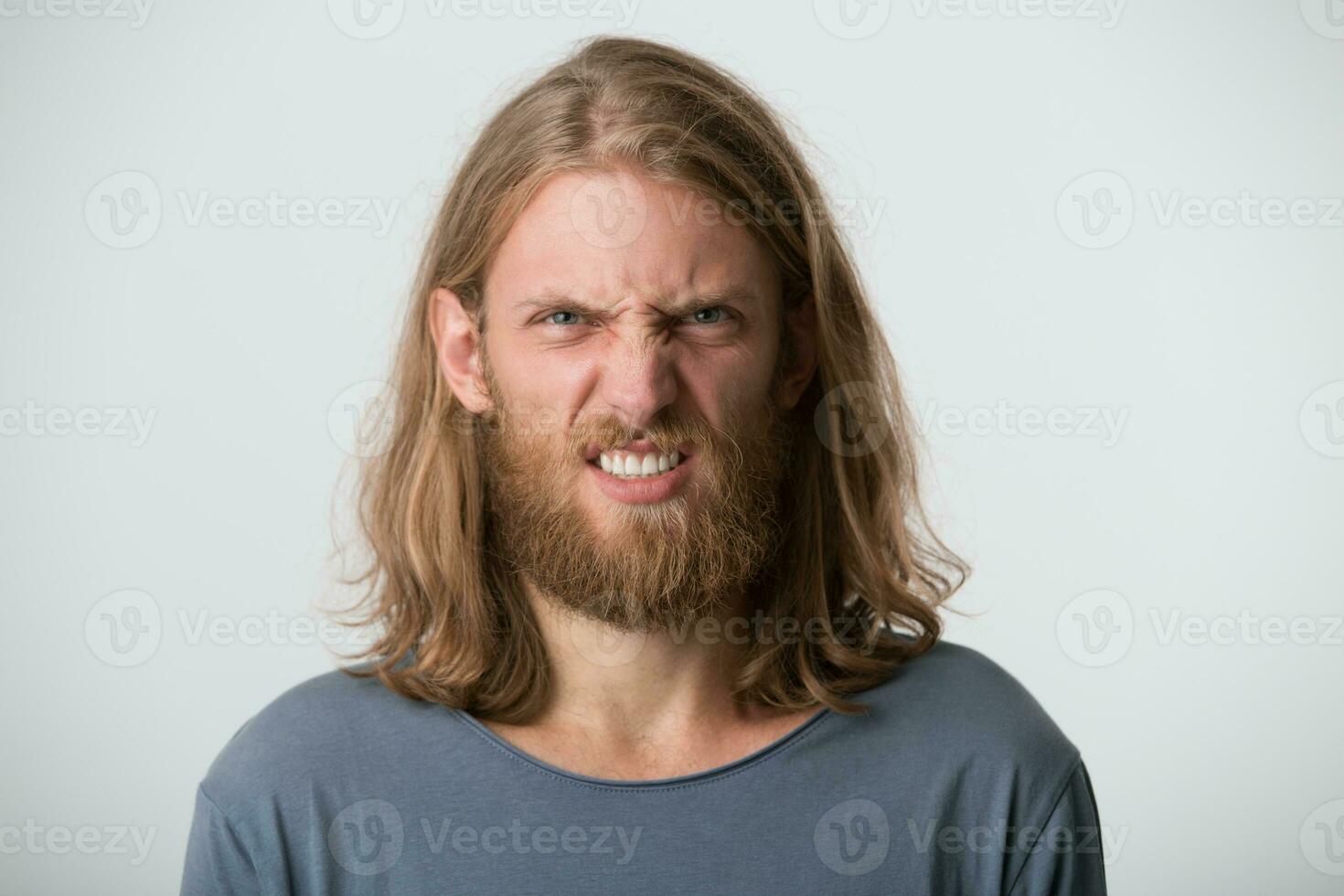 Closeup of mad crazy young man with beard and blonde long hair wears gray t shirt looks angry and displeased isolated over white background photo