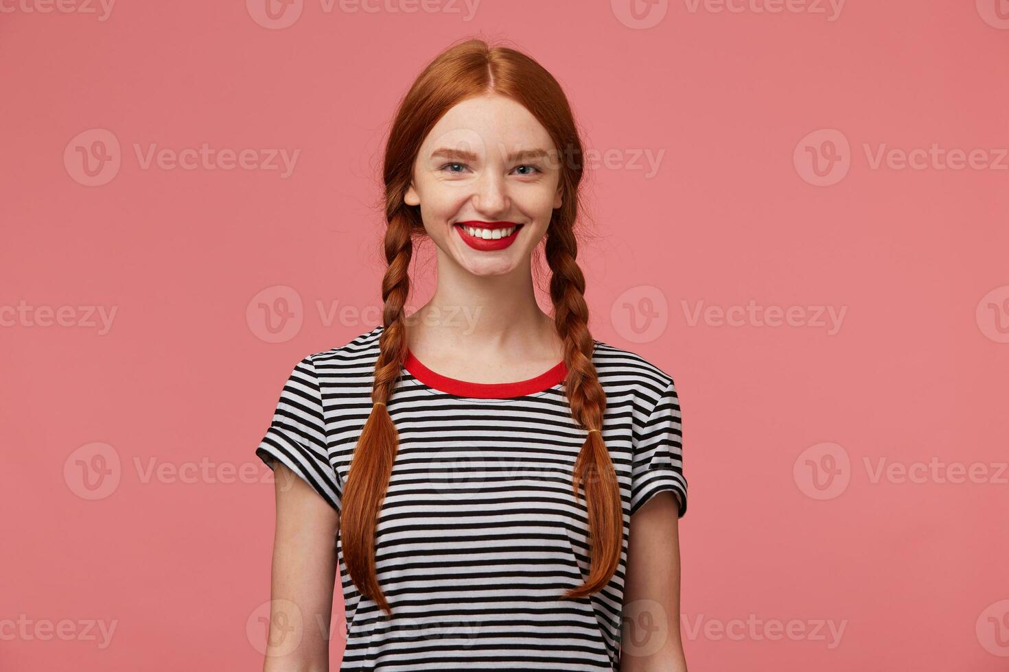 bonito bonito Pelirrojo niña con rojo labios, dos trenzas, encantador sonrisa, muestra blanco sano dientes, vestido en despojado camiseta, aislado en un rosado antecedentes foto