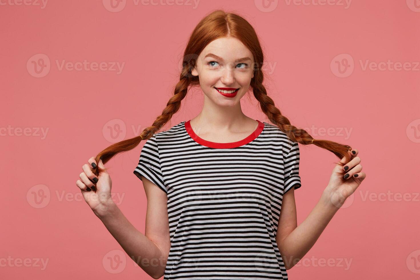Nice pretty red-haired girl with red lips holding two braids in hands dressed in stripped t-shirt, flirting playfully looking at the left upper corner plotting adventure isolated on a pink background photo