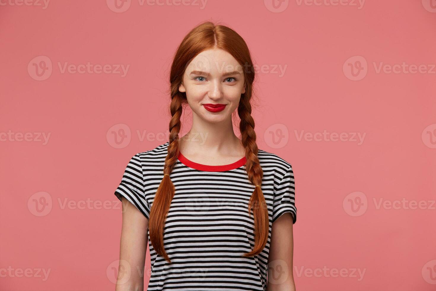 retrato de encantador bonito hermosa niña con Pelirrojo trenzas rojo labios, bonito sonrisa, vestido en despojado camiseta, aislado en un rosado antecedentes foto