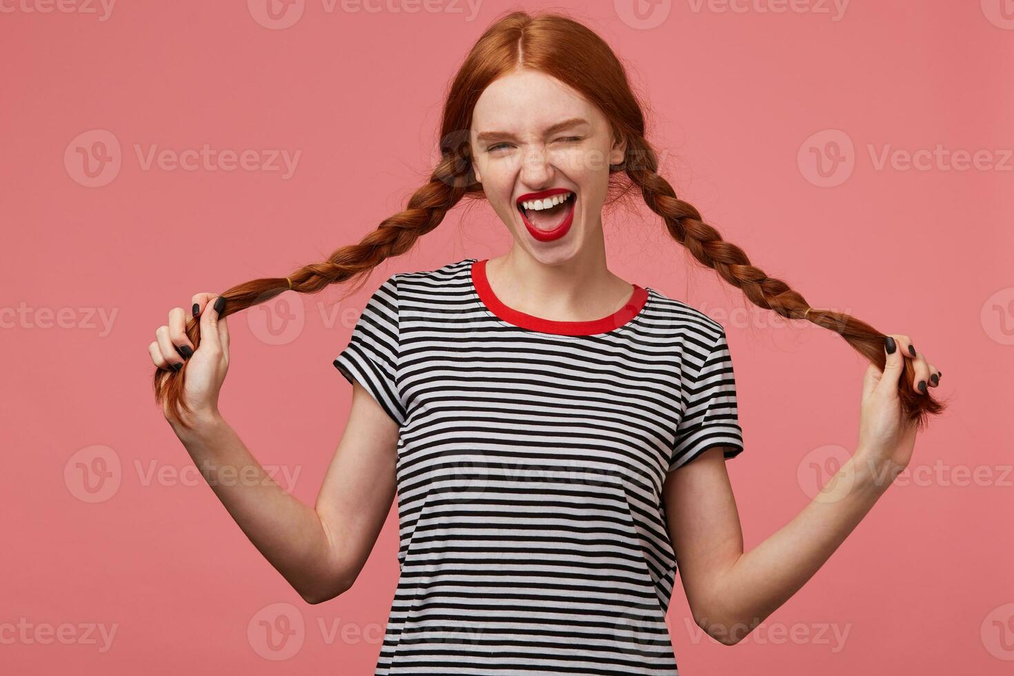 Funny joyful girl holding two red haired braids in hands, with red lips, dressed in stripped t-shirt, dressed in stripped t-shirt, winks happy playfully looking camera isolated on a pink background photo