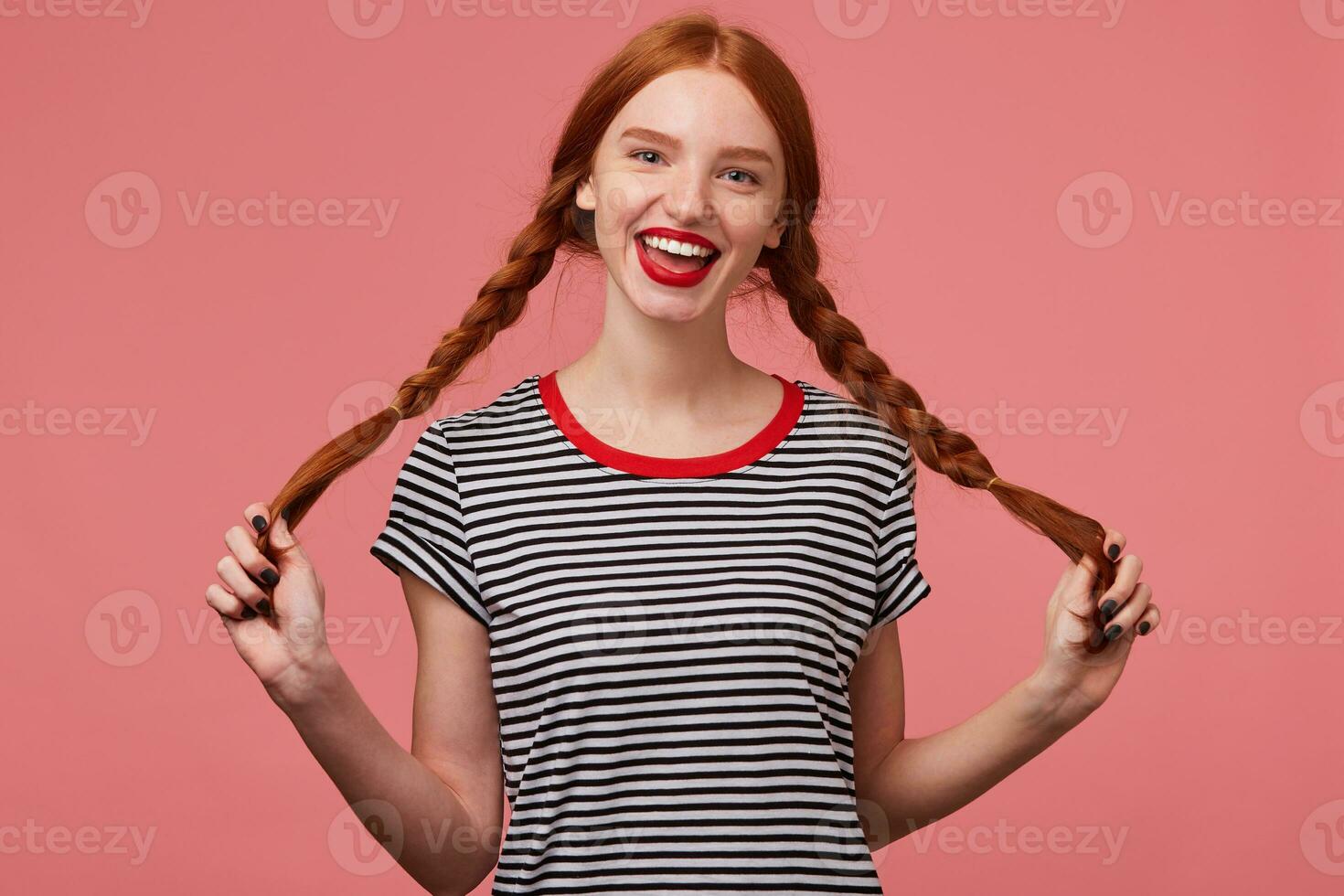 encantador niña participación dos rojo peludo trenzas en manos, con rojo labios, vestido en despojado camiseta, vestido en despojado camiseta, contento en broma mirando cámara aislado en un rosado antecedentes foto