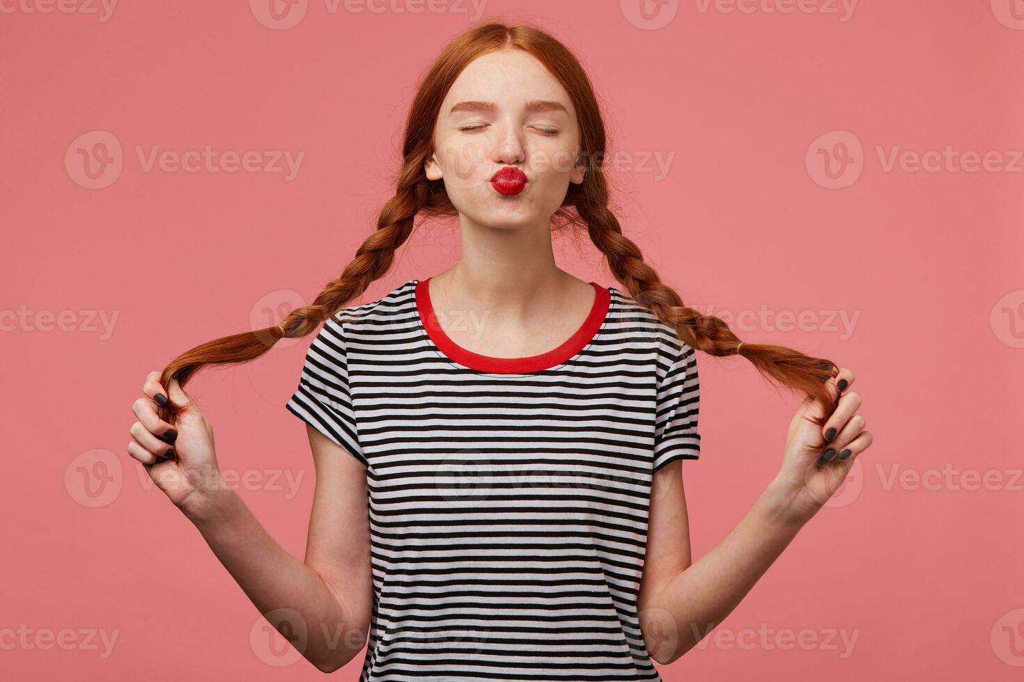 Sweet red-haired girl with red lips sends air kiss to a camera with closed eyes holding two braids in hands dressed in stripped t-shirt, flirting isolated on a pink background photo