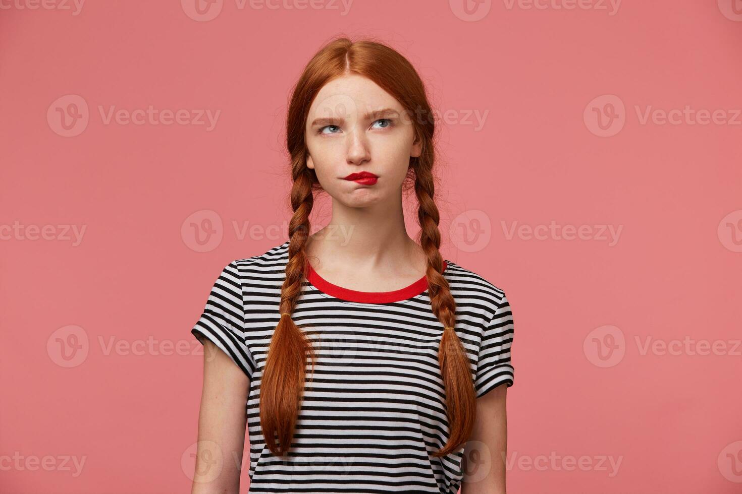 Thoughtful girl with two red haired braids biting red lip doubts about something, dressed in stripped t-shirt, looks to the upper right corner isolated on pink background photo