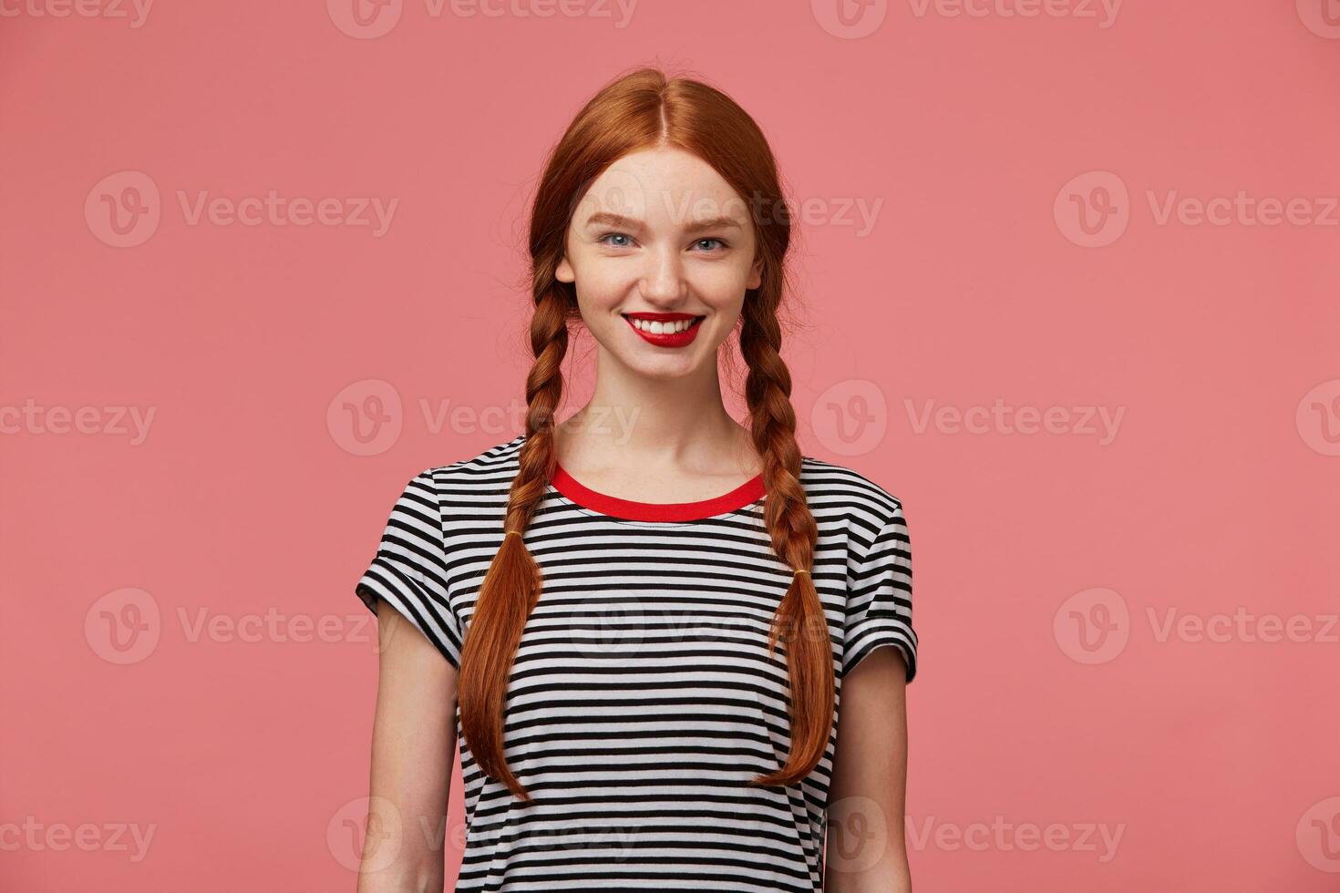 atractivo hermosa reconfortante bonito Pelirrojo niña con rojo labios, dos trenzas, encantador sonrisa, muestra blanco sano dientes, vestido en despojado camiseta, aislado en un rosado antecedentes foto