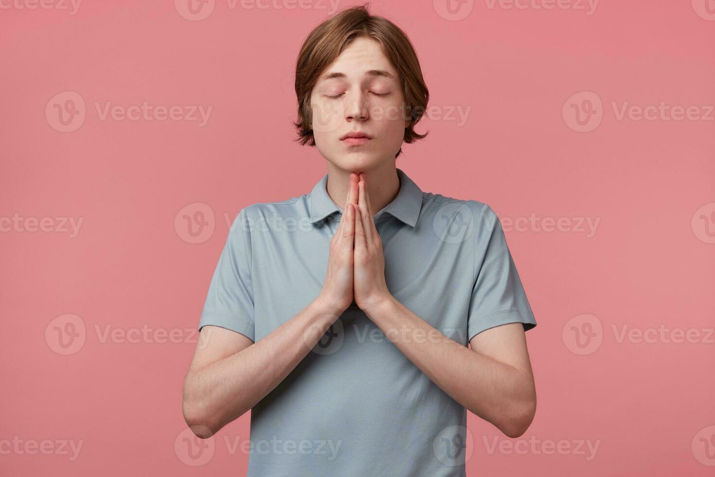 Kind obedient guy praying hands clasped hoping for best asking for forgiveness, pray with your eyes closed, over pink background. Human emotion facial expression feeling photo