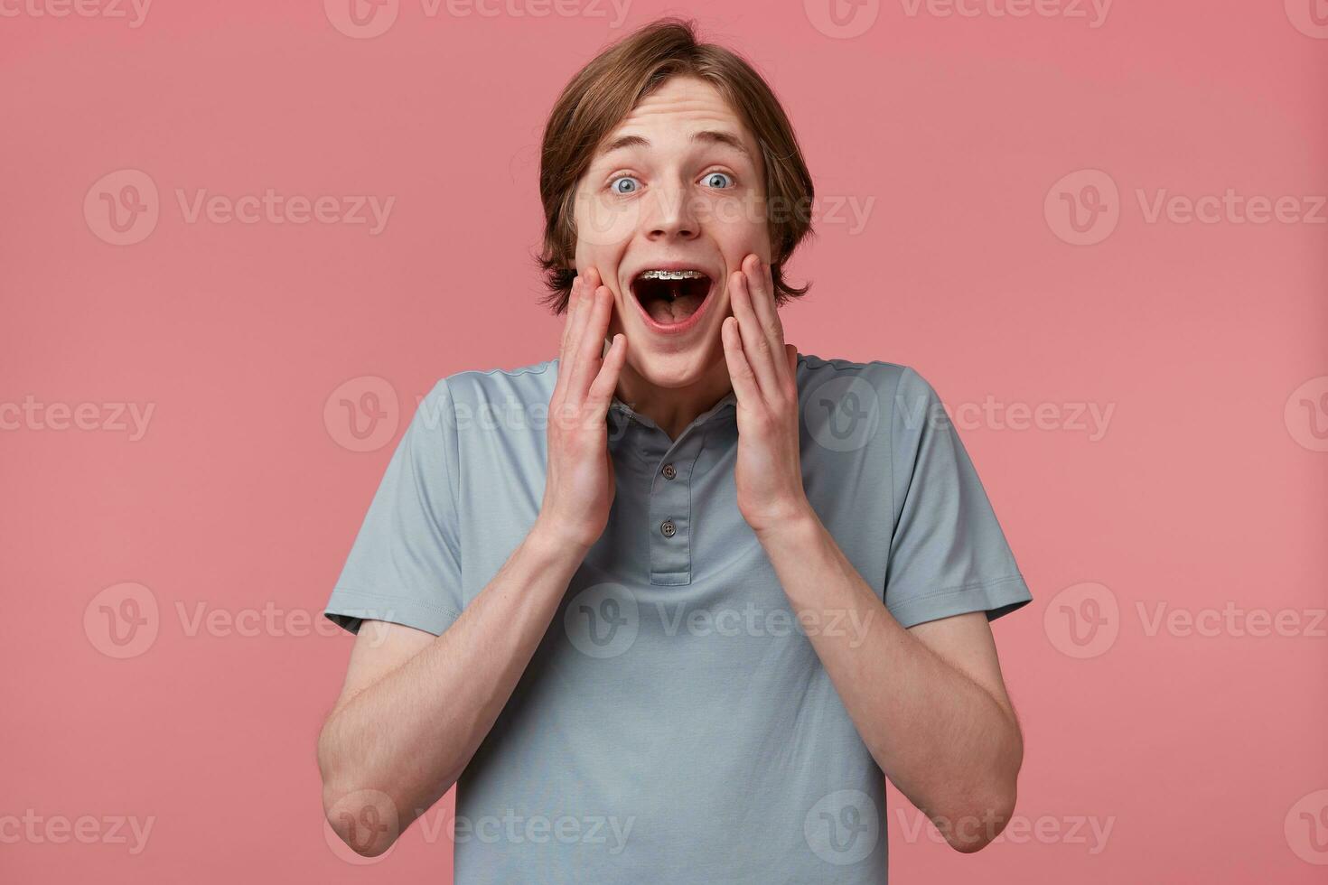 Amazed excited young guy touching his face can't believe in his luck, with long neatly combed hair and braces on teeth wears polo t-shirt shouting and feels happy surprised isolated over pink wall photo