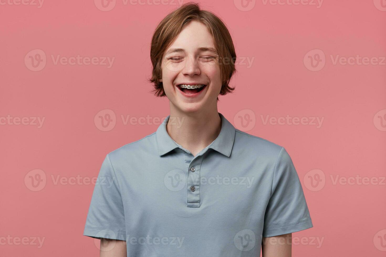 Close up of nice blue-eyed neatly combed young guy with braces on teeth joyfully laughing closed his eyes of fun wears polo t-shirt looks happy isolated over pink background photo