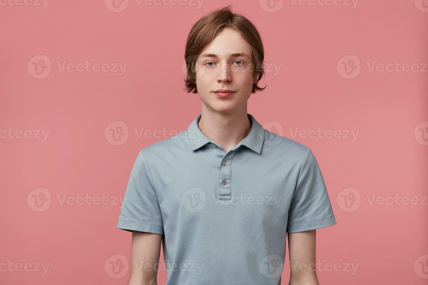 Young man did not slept for a long time, tired, zombie eyes, slightly-open eyes, looks calm, express no feelings, dressed in polo T-shirt, isolated over pink background photo