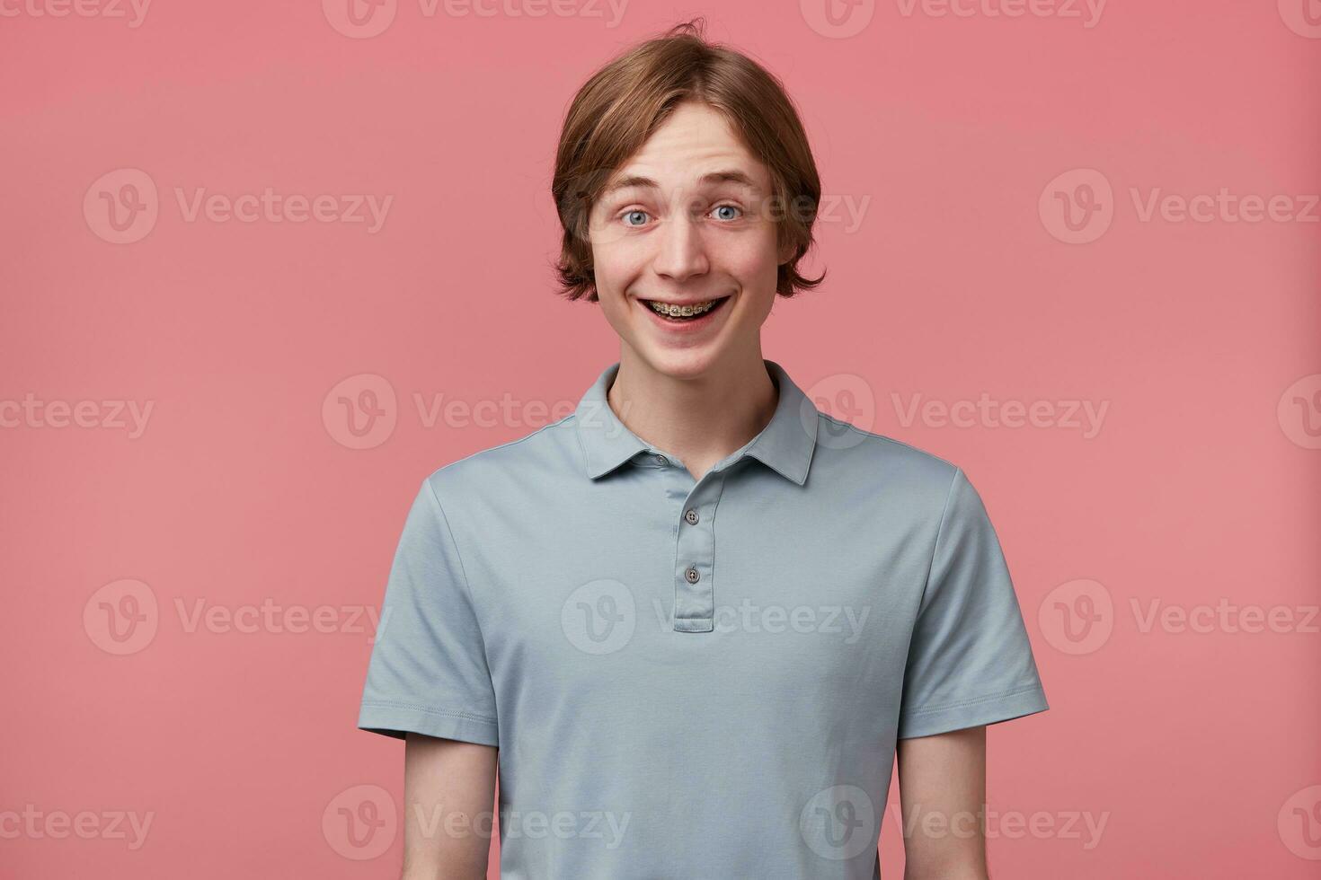 Portrait of excited surprised glad blue-eyed neatly combed young guy with braces on teeth wears polo t-shirt happily smiles and feels happy isolated over pink background photo