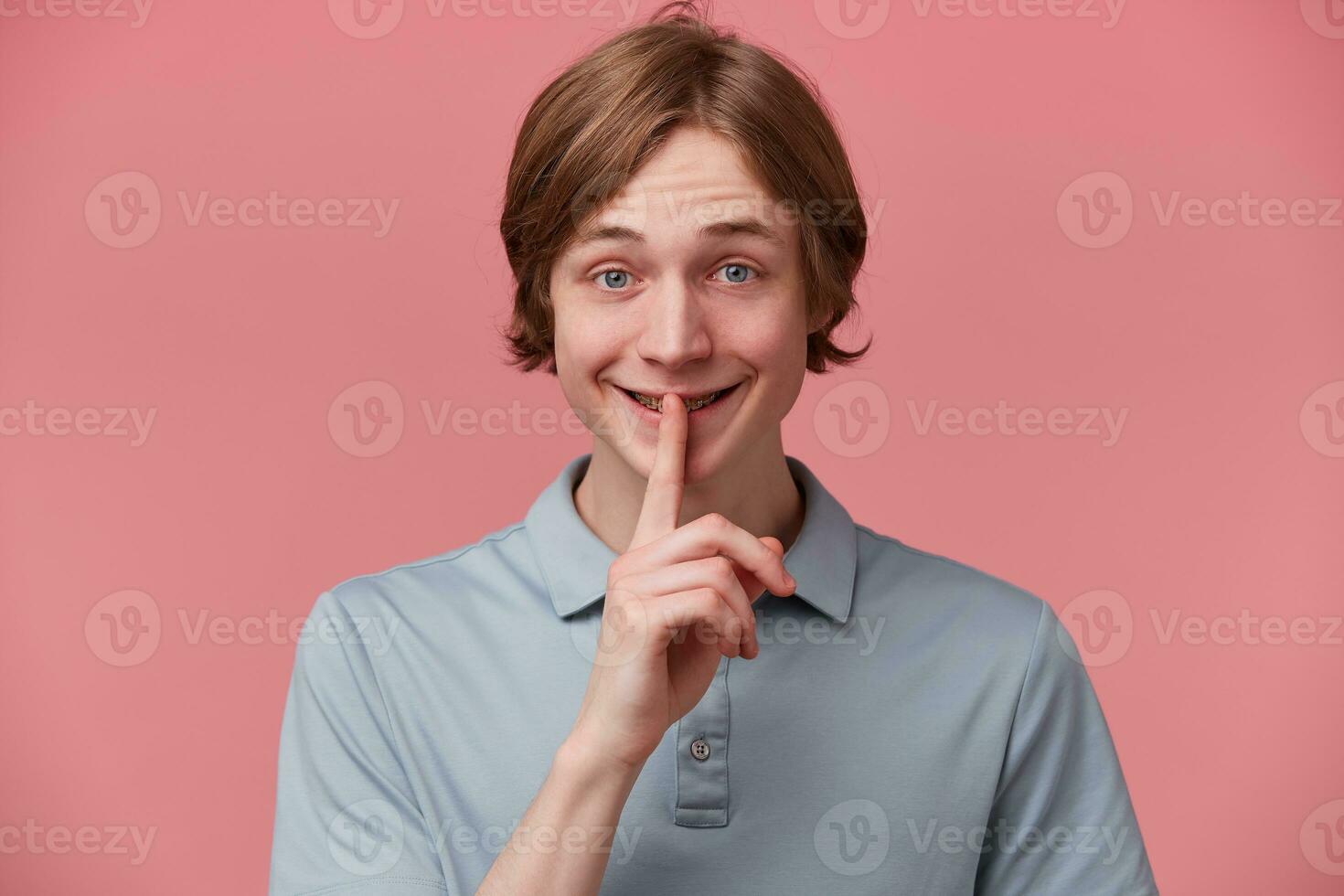 Happy smiling young guy keeps fore finger on lips, calls to keep a secret, do not tell anyone, keep quiet, do not make noise, demonstrates silence gesture, isolated over pink background photo