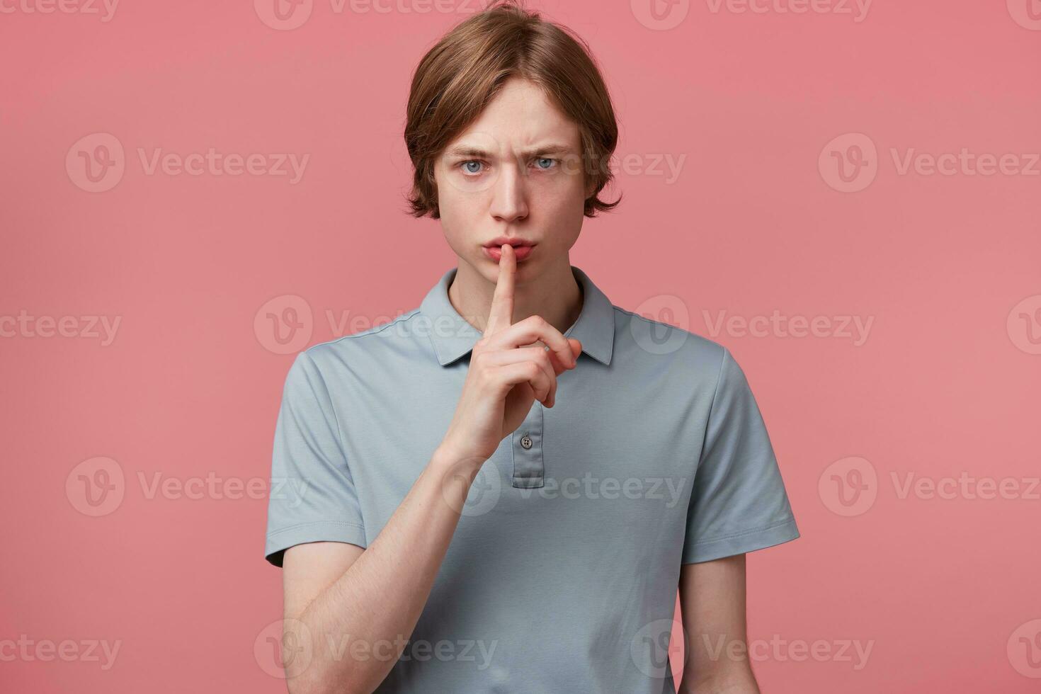 Young serious frowned guy keeps fore finger on lips, calls to keep a secret, do not tell anyone, keep quiet, do not make noise, demonstrates silence gesture, isolated over pink background photo