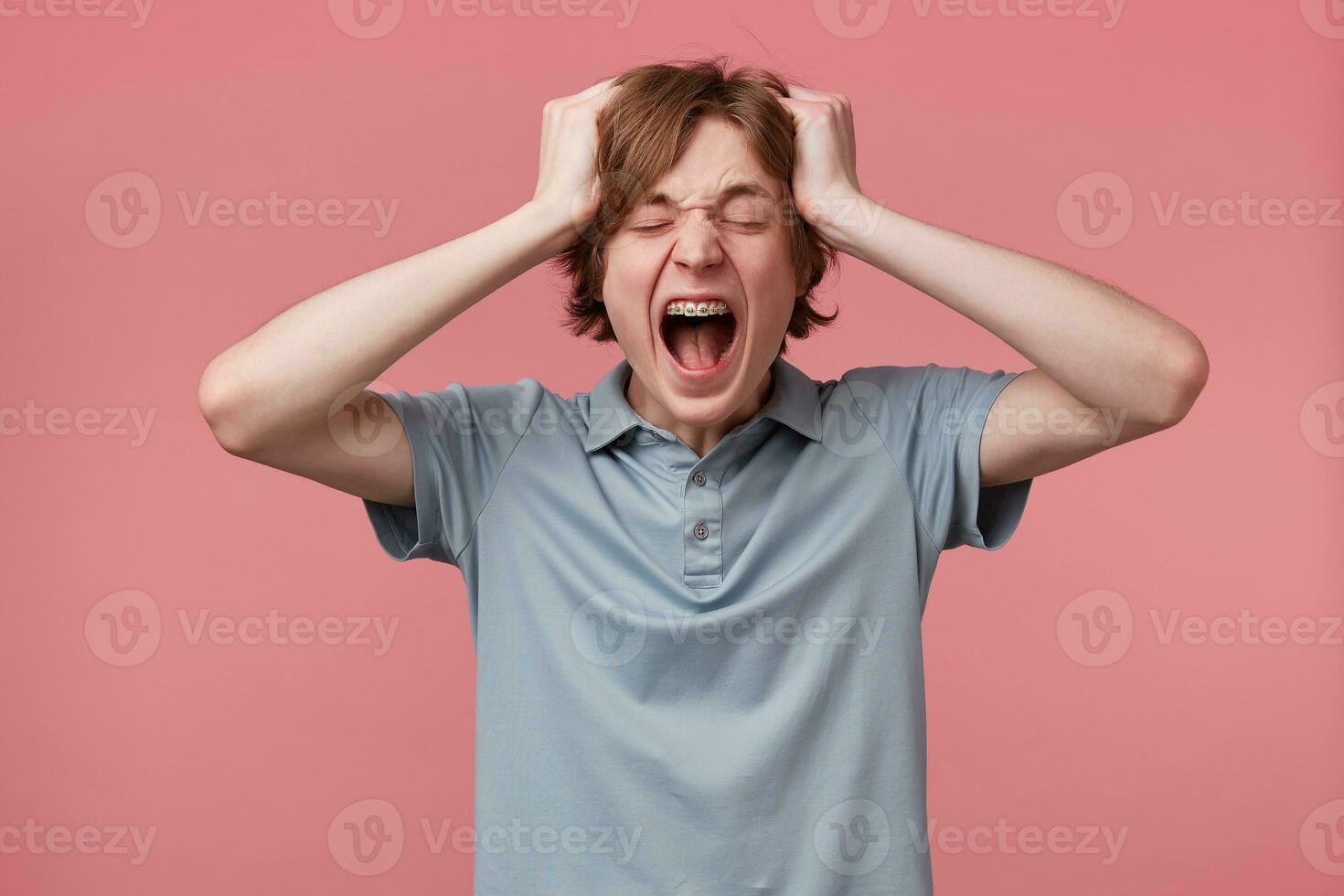 With young man happened something terrific, keeps both hands on head, shouts loudly with wide opened mouth, eyes closed, over pink background. People, negative reaction and feelings concept. photo