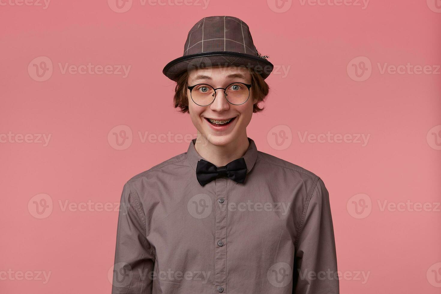 Surprised excited attractive young guy smartly dressed in shirt hat and black bowtie wears glasses has orthodontic brackets, nicely widely smiling isolated on pink background photo