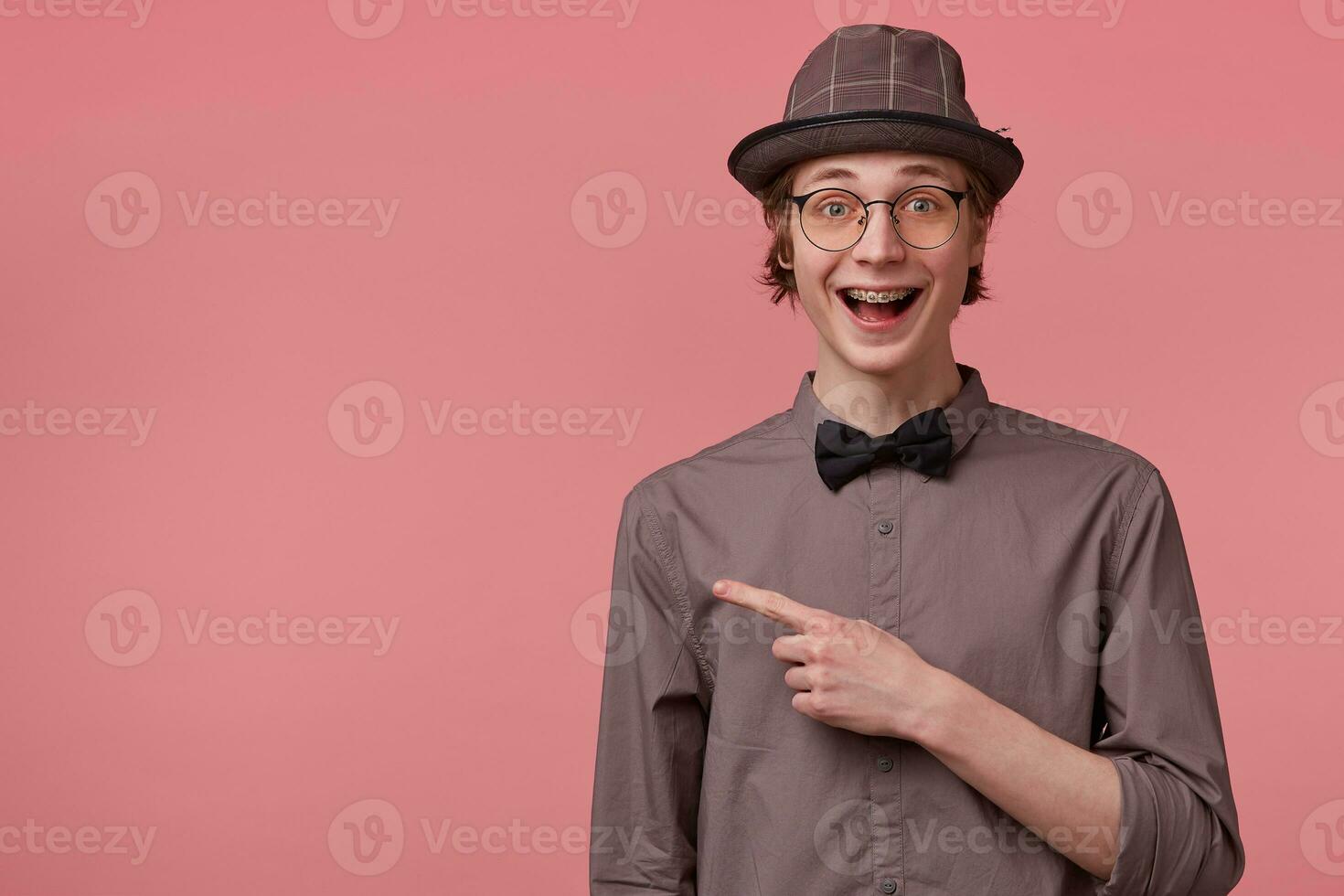Elegant young guy opened his mouth in excitement, is overwhelmed with positive emotions happiness joy isolated on pink, pointing index finger to the left on copy space photo