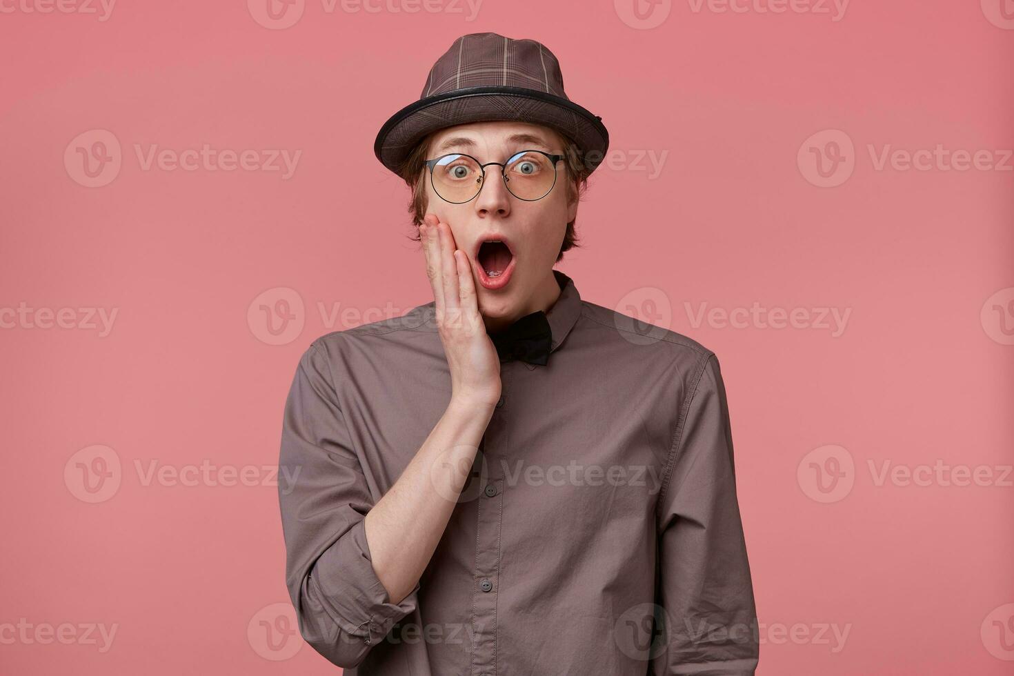 Young guy seriously shocked very amazed with opened mouth touching face smartly dressed in shirt hat and black bowtie wears glasses feels surprised isolated on pink background photo