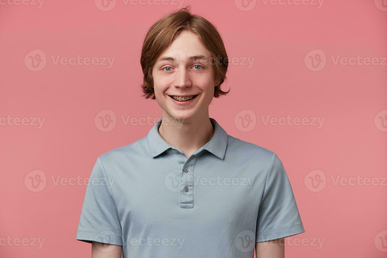 Close up of blue-eyed neatly combed young guy with braces on teeth wears polo t-shirt slightly smiles and feels happy isolated over pink background photo