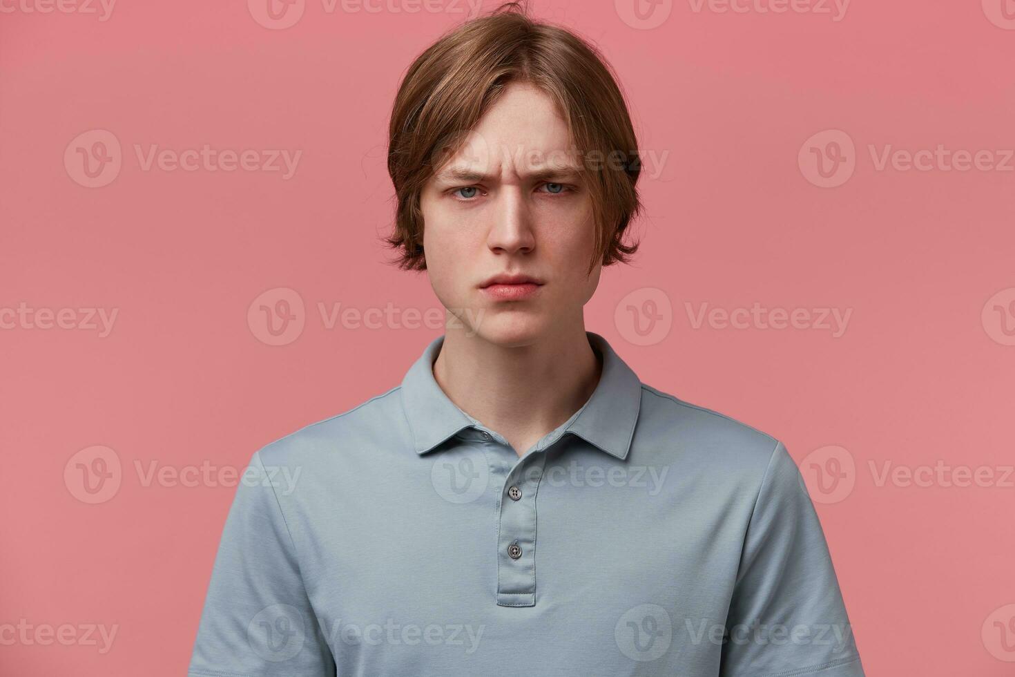 Close up of angry furious unsatisfied neatly combed young guy, eyebrows frowned, eyes aggressive, dressed in casual polo T-shirt, isolated over pink background photo
