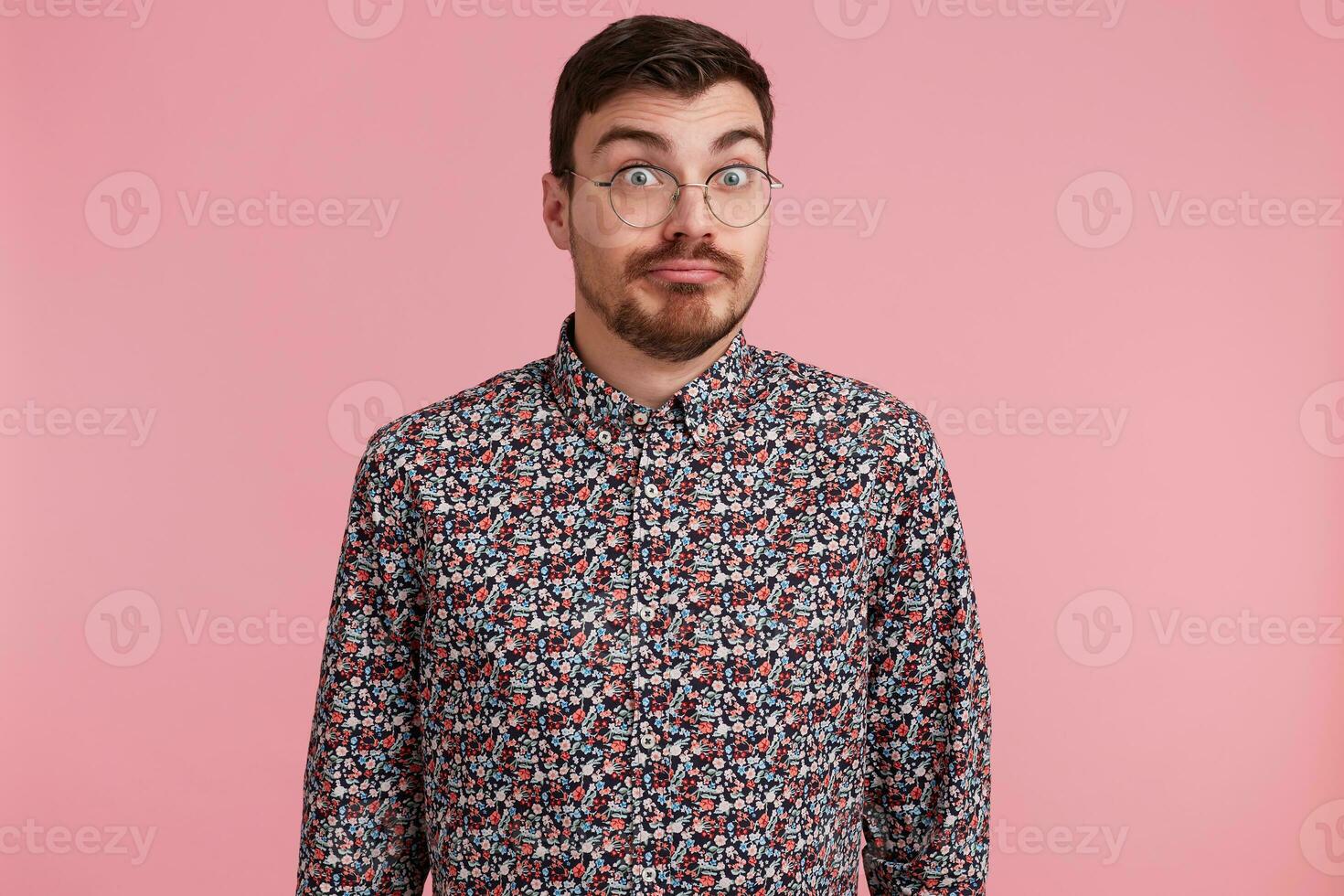 Unsure uncertain surprised man stares through spectacles wearing colorful shirt shrug shoulders in uncertainty, over pink background photo